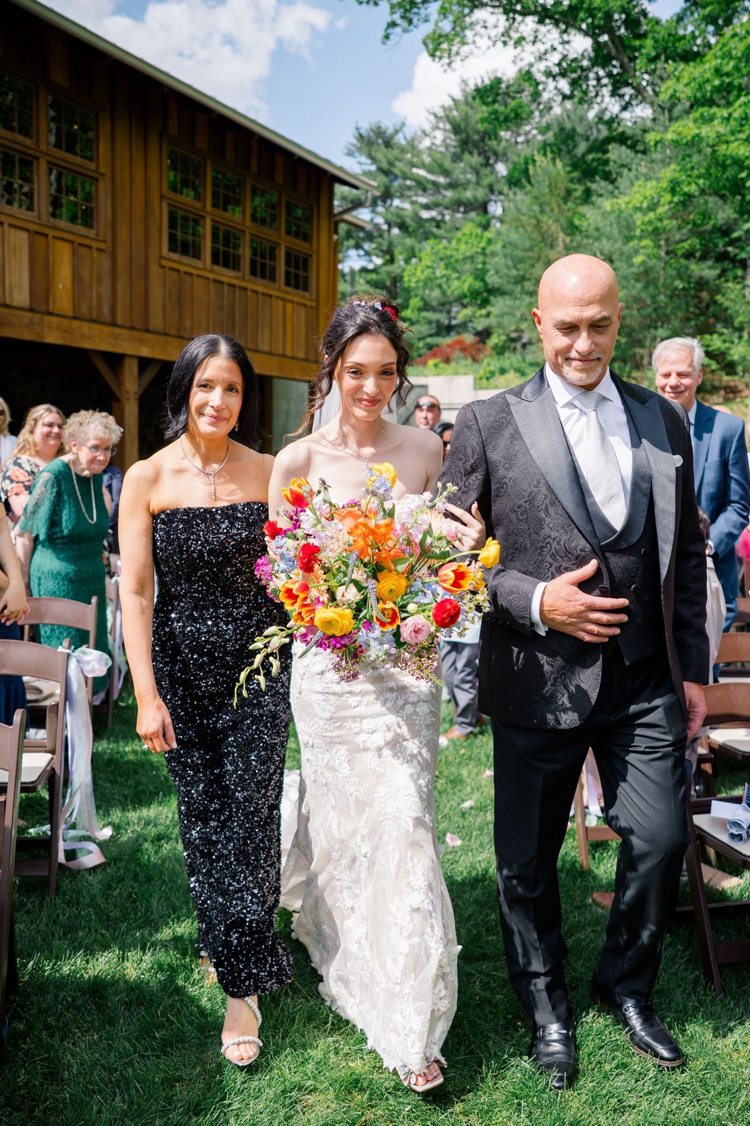 Bride in lace dress walking down aisle with orange bouquet, escorted by wedding party members.