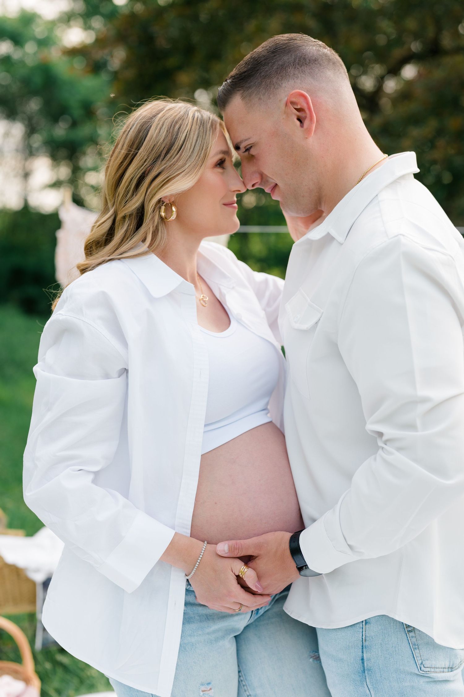 Intimate pregnancy photoshoot moment in white outfits against natural outdoor background.