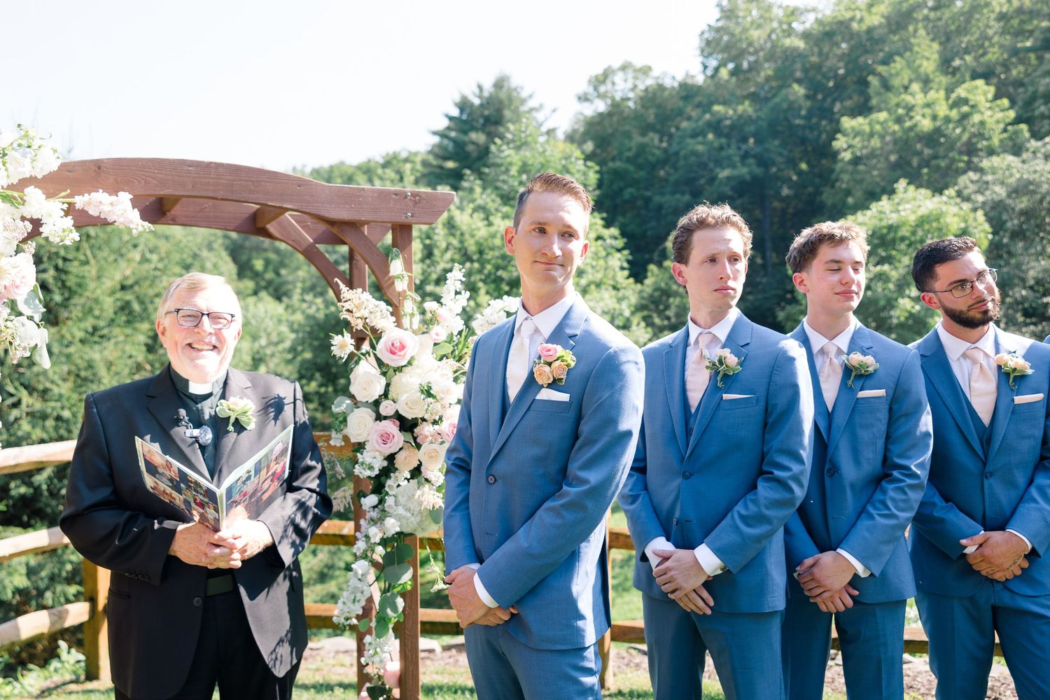 Wedding party stands at outdoor ceremony with blue suits and floral arrangements.