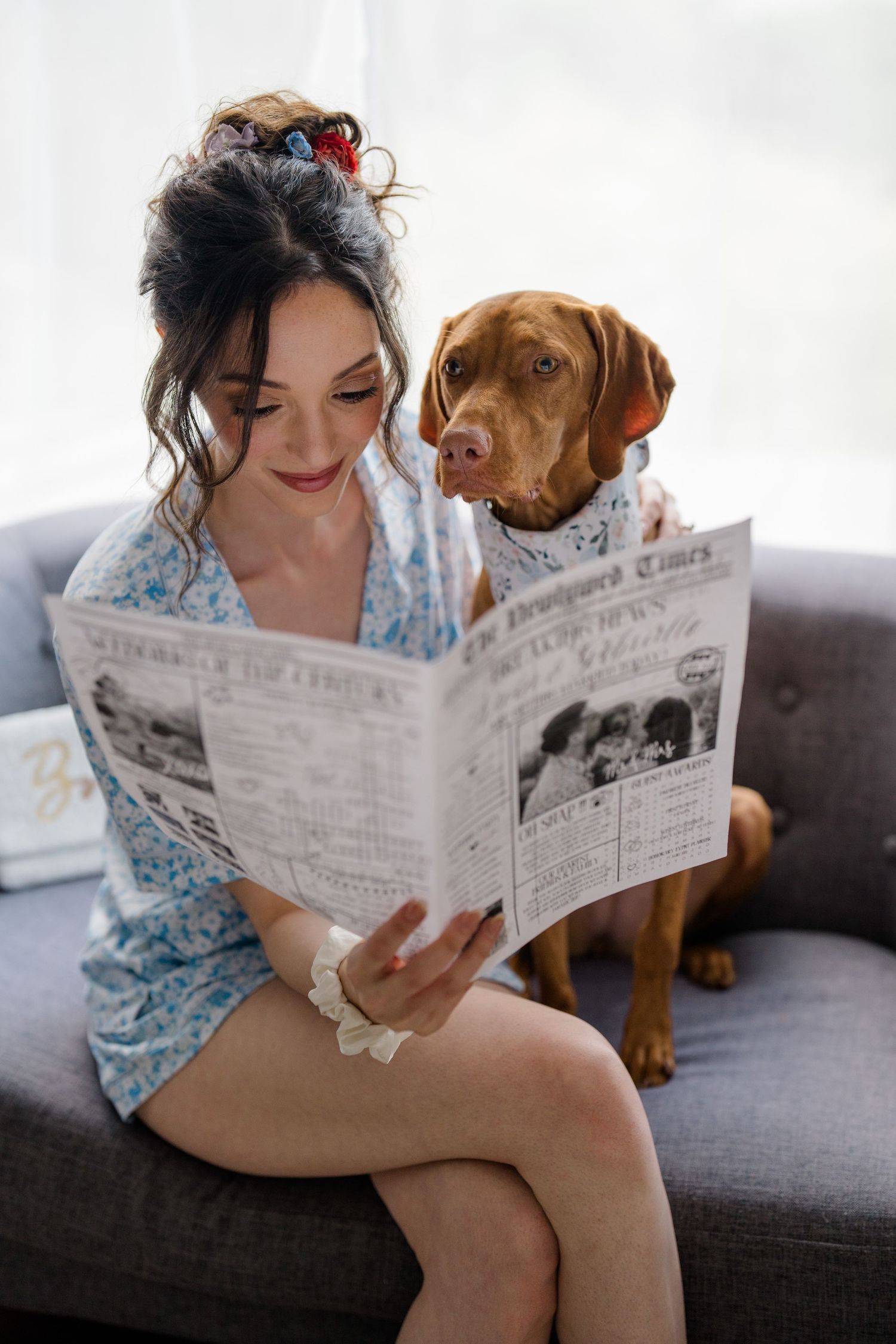 A rustic comfort scene shows a woman and brown dog reading a newspaper together on a dark gray couch.