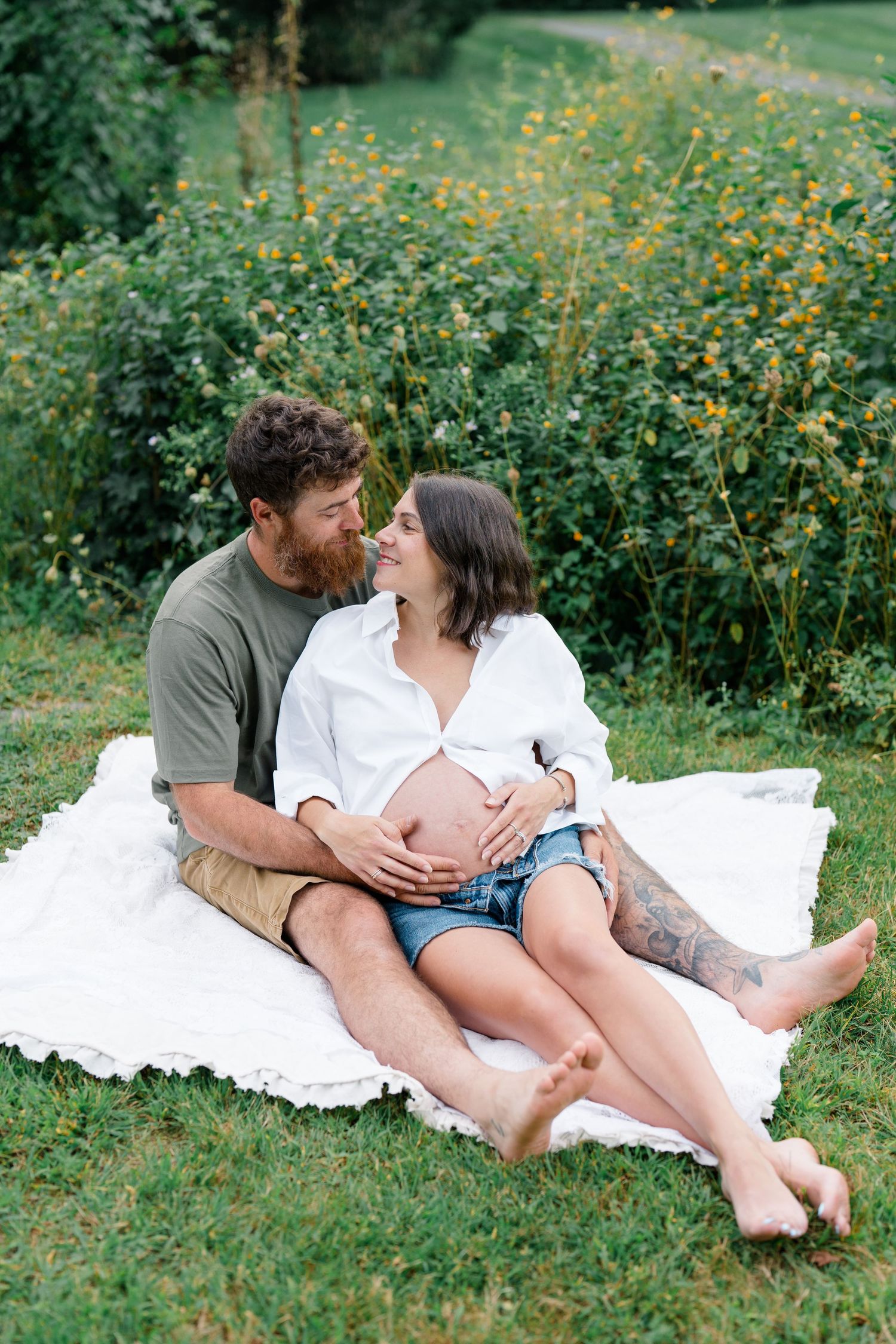 Couple sitting on white blanket outdoors, man embracing pregnant woman's belly, surrounded by lush green garden.