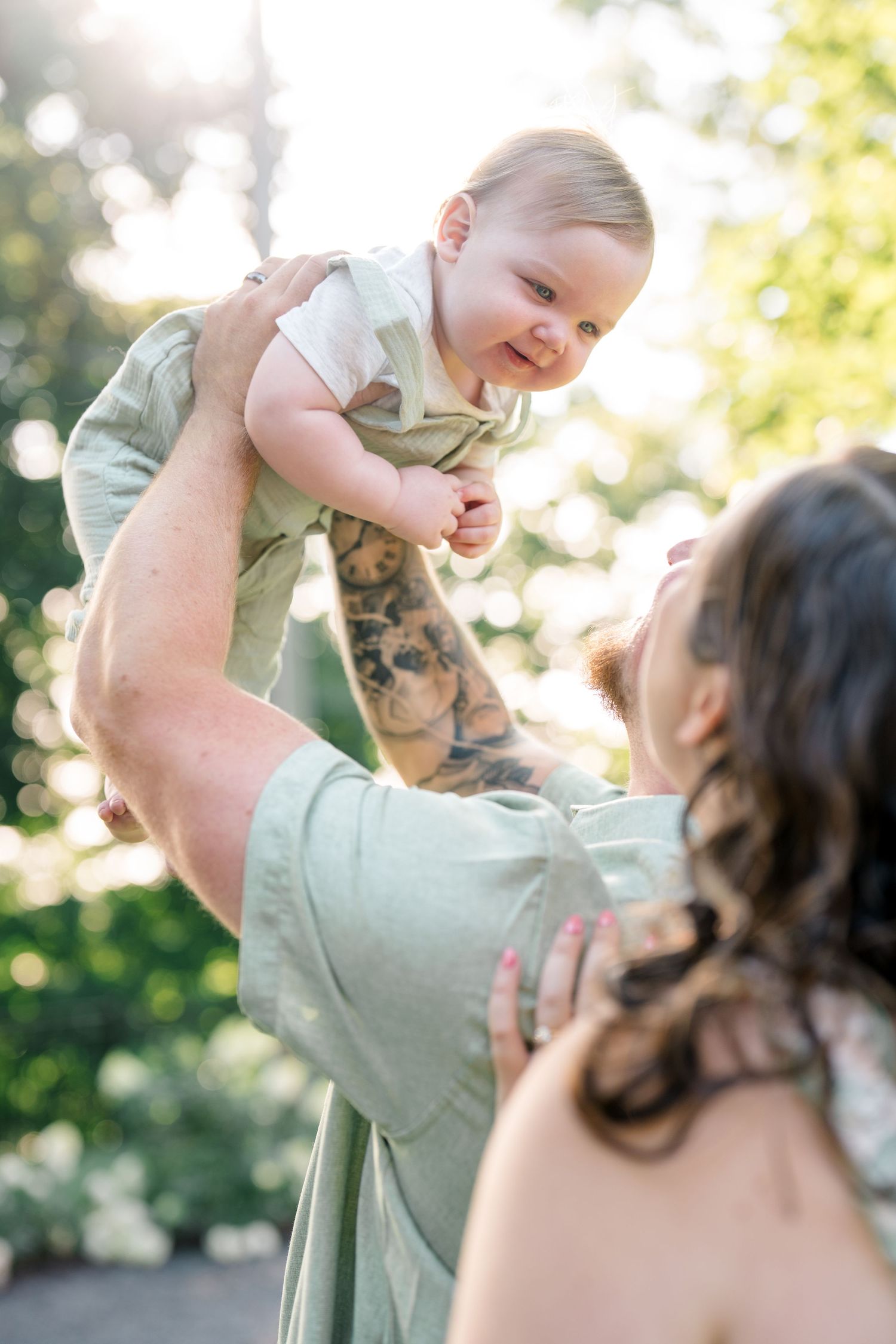 Tattooed parent joyfully lifting smiling baby outdoors in sunlit garden, with another parent watching nearby.