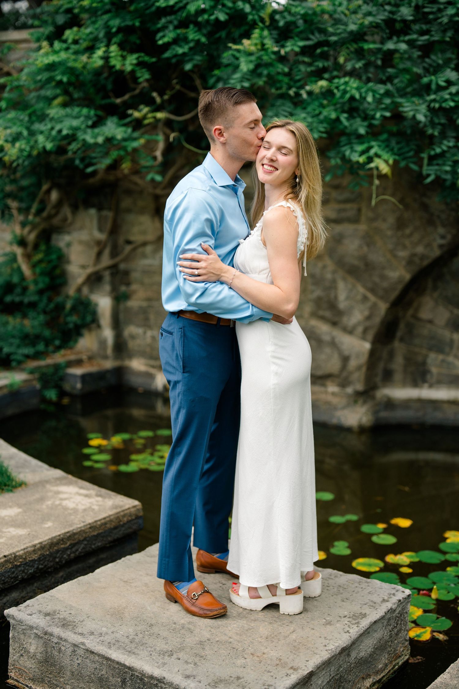 A couple embraces on a stone platform by a lily pond, the man kissing her cheek as she smiles in a white dress.