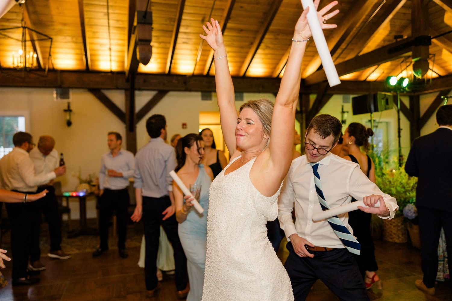 Bride joyfully dancing with arms raised at indoor wedding reception while guests celebrate around her in rustic venue.