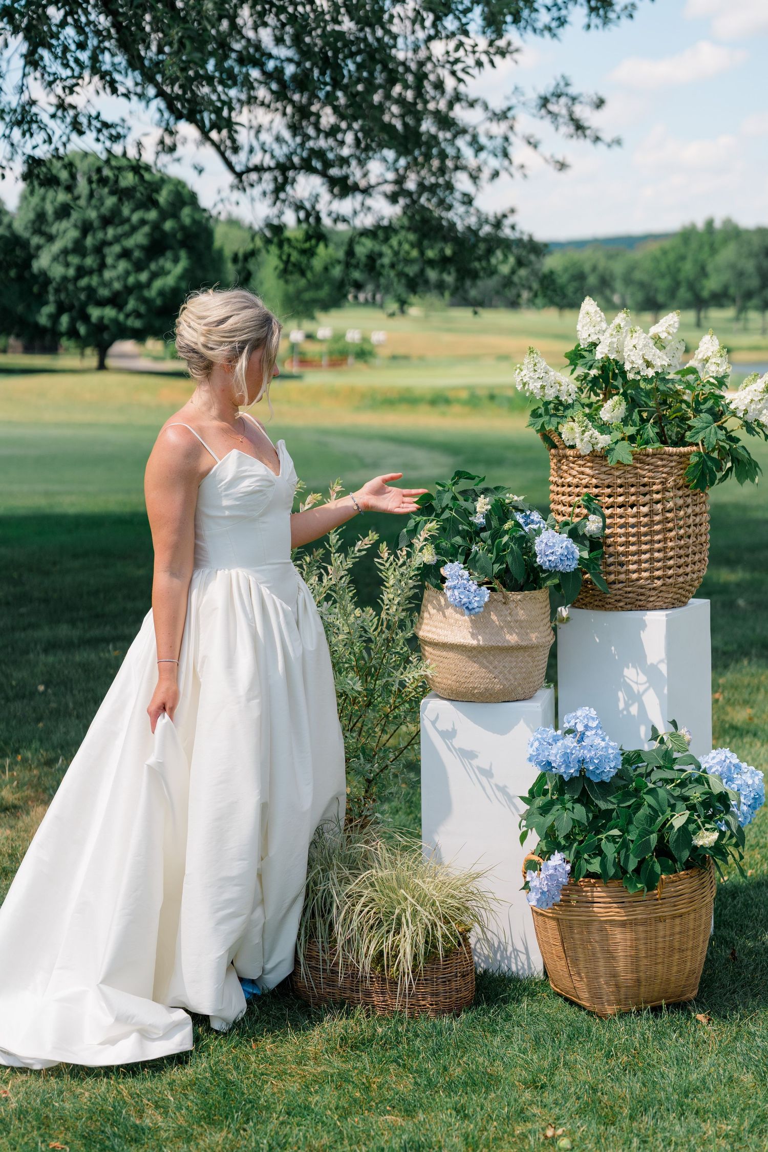 Elegant bride in a white spaghetti-strap ball gown admires floral arrangements in wicker baskets on white pedestals.
