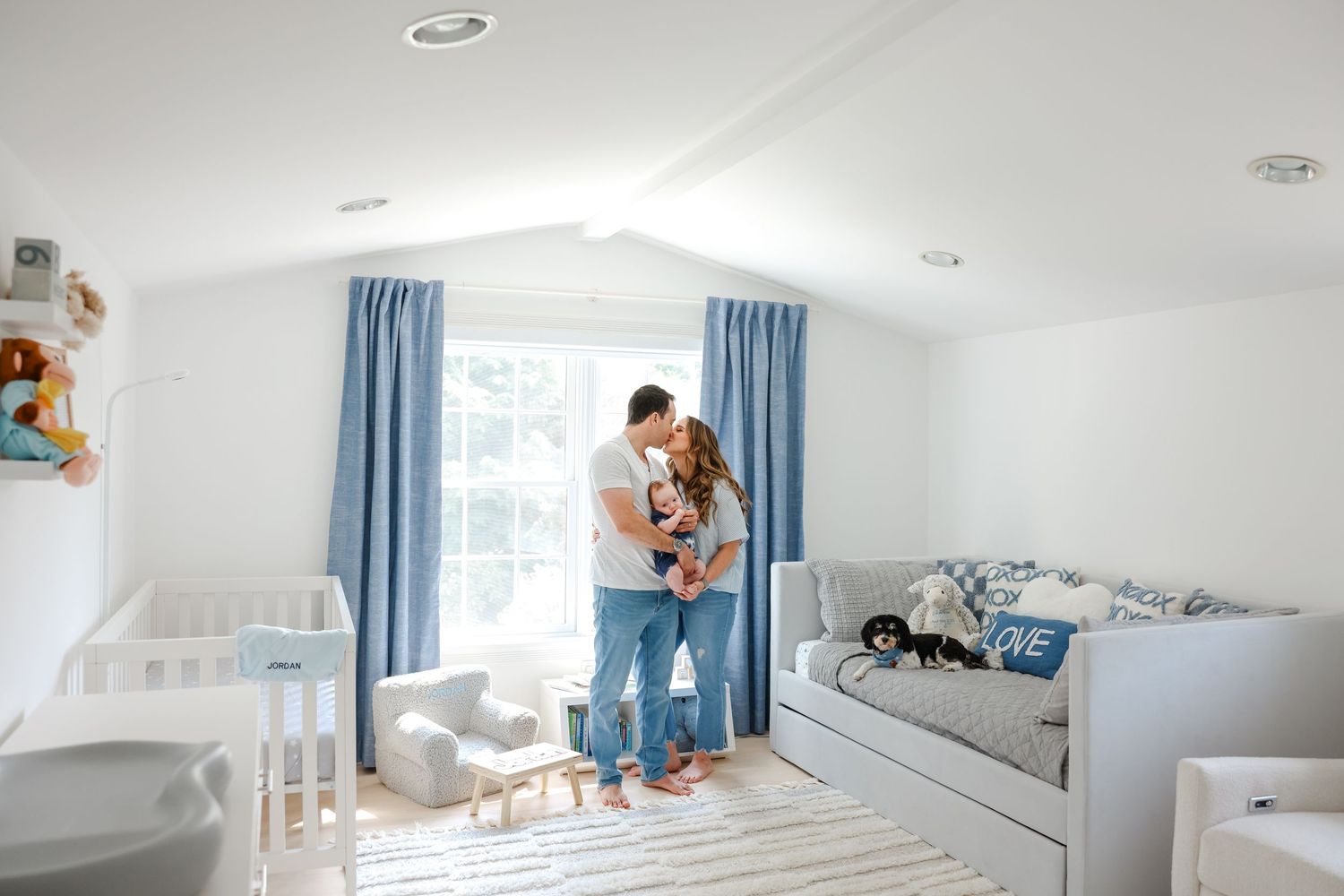 A bright nursery room decorated in blue and white with curtains, a crib, and sofa near a large window.