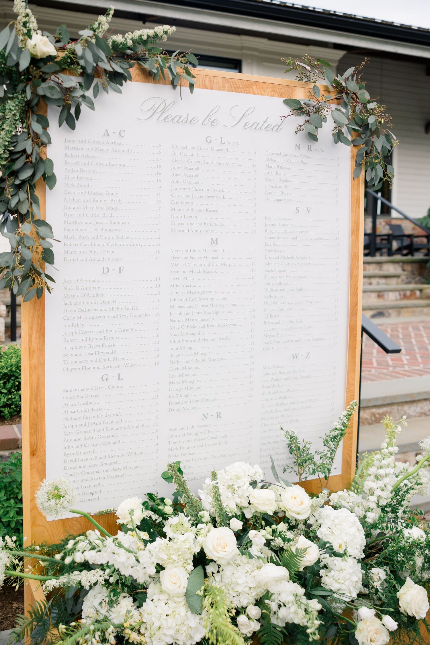 Wedding seating chart display decorated with eucalyptus and white flowers in a wooden frame.