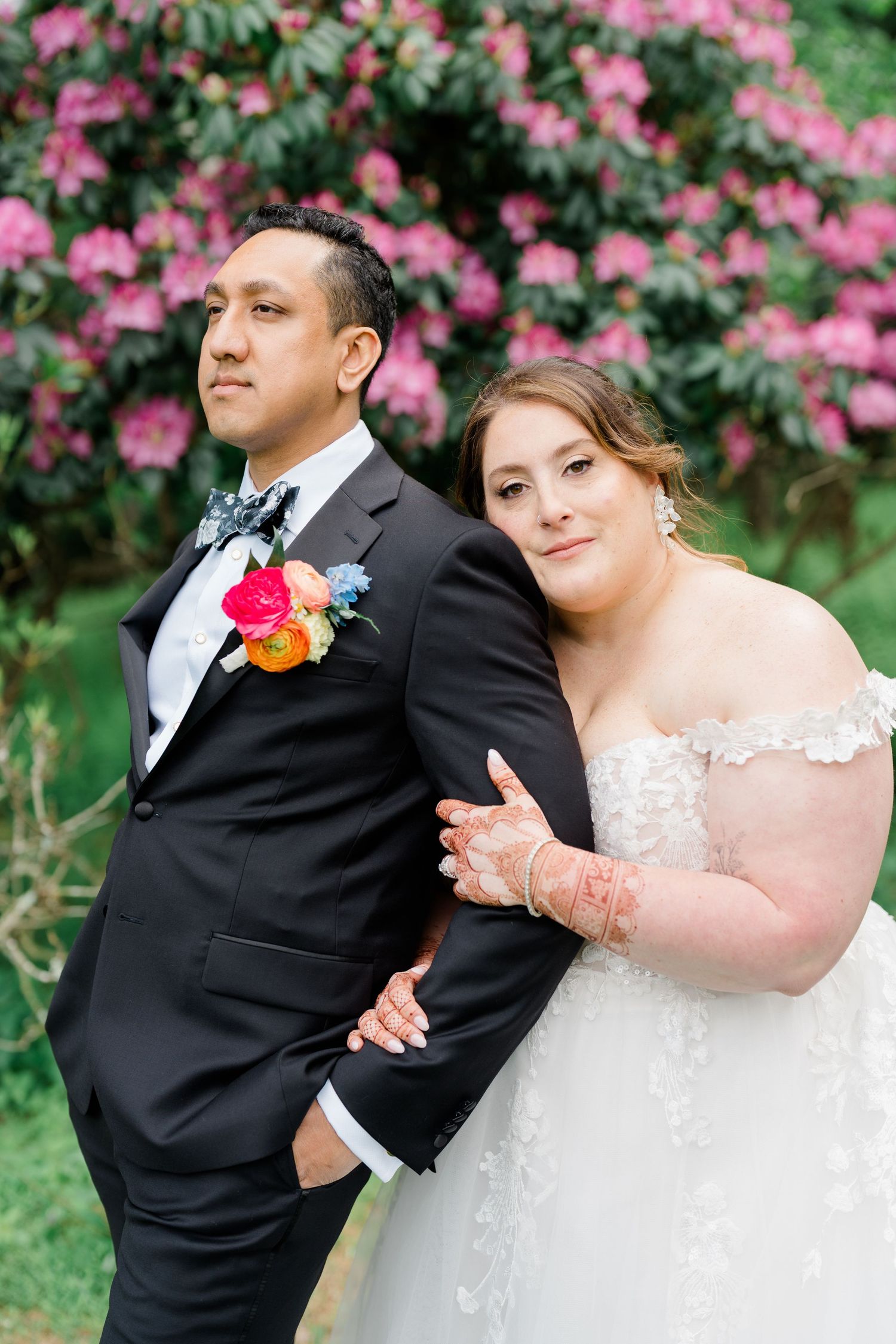 A bride and groom pose together outdoors with pink azalea bushes blooming behind them.