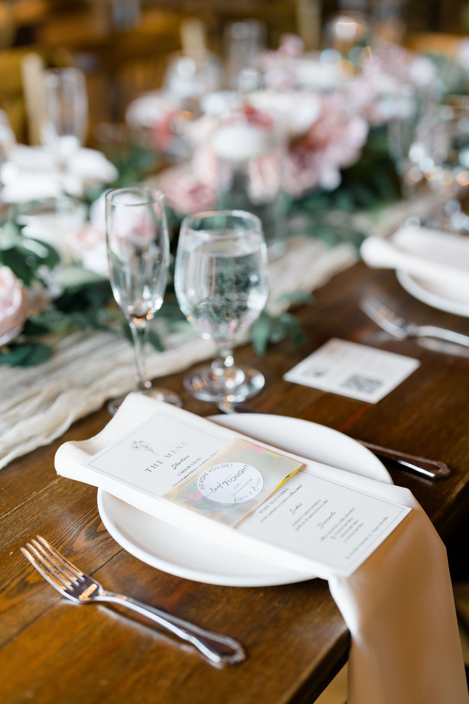 Wedding reception table setting with menu cards, crystal glasses, and white plates on rustic wooden table.
