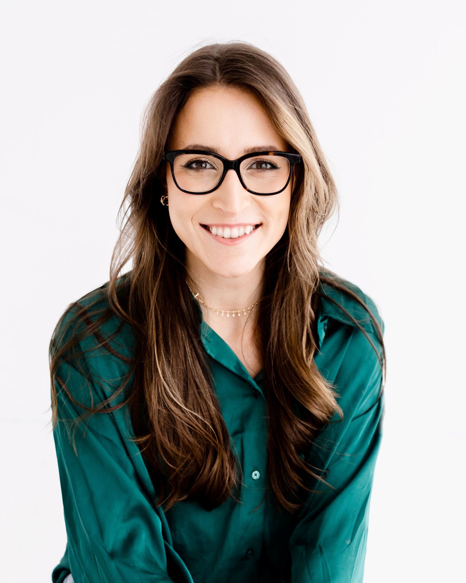 Smiling young woman with long brown hair and black glasses wearing a teal silk blouse against white background.