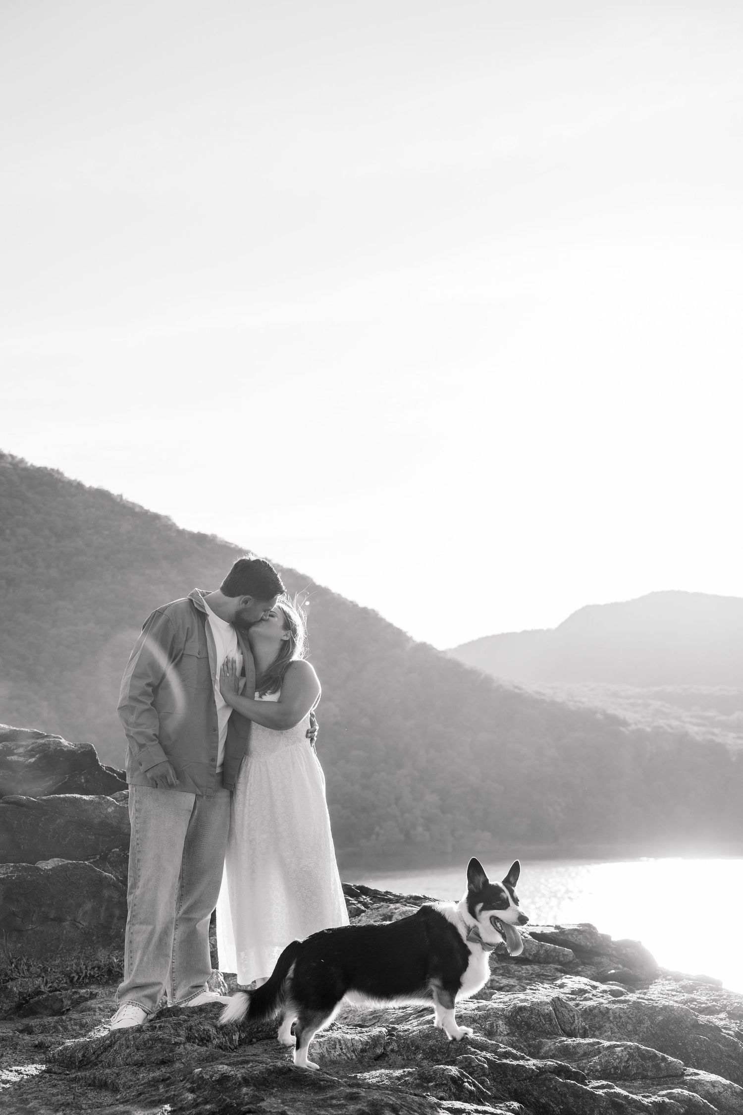 Black and white photo of a couple embracing on rocky shore with a Corgi dog, mountains and lake in background.