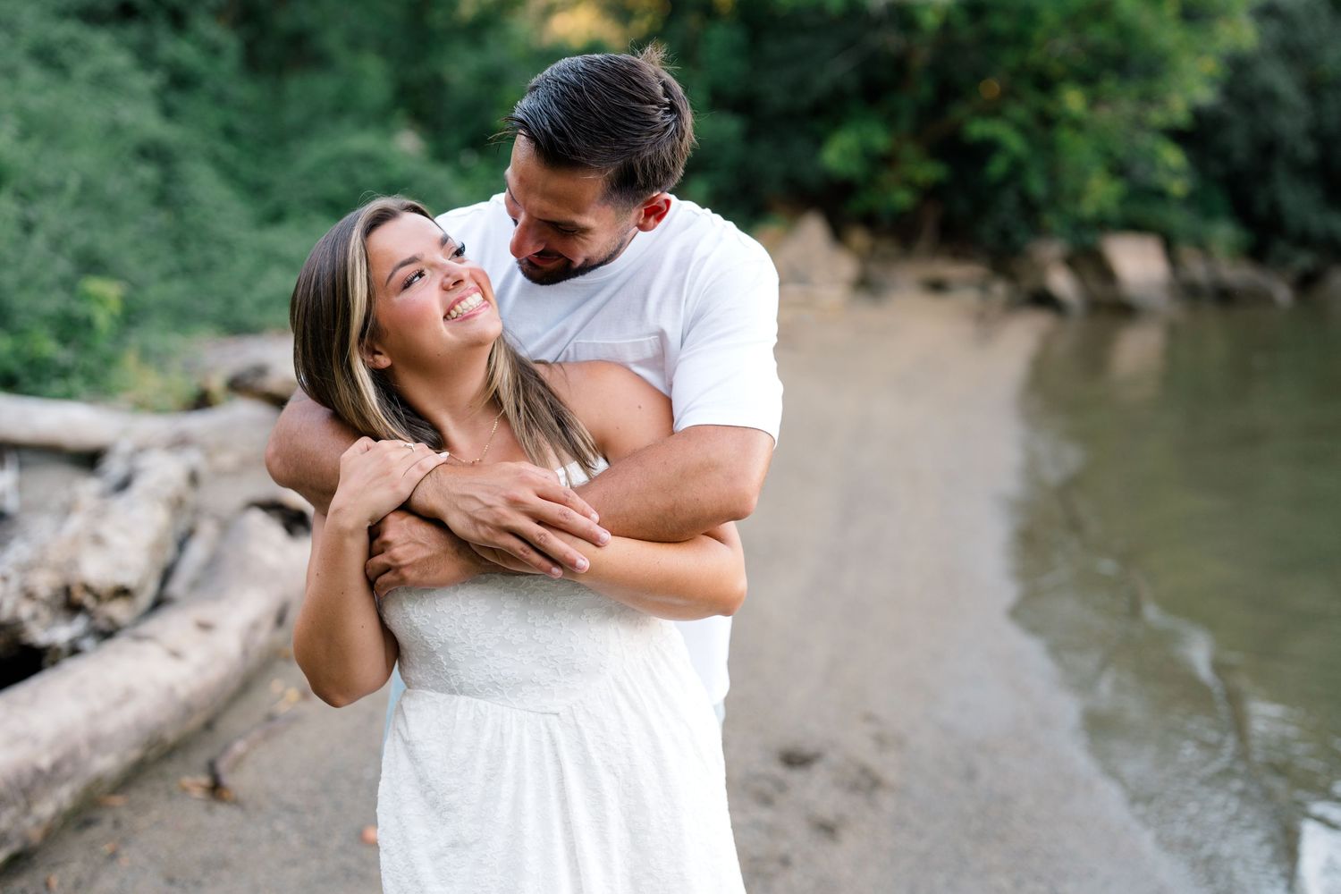 Couple embracing and smiling on a rocky beach shore surrounded by lush green trees and driftwood.