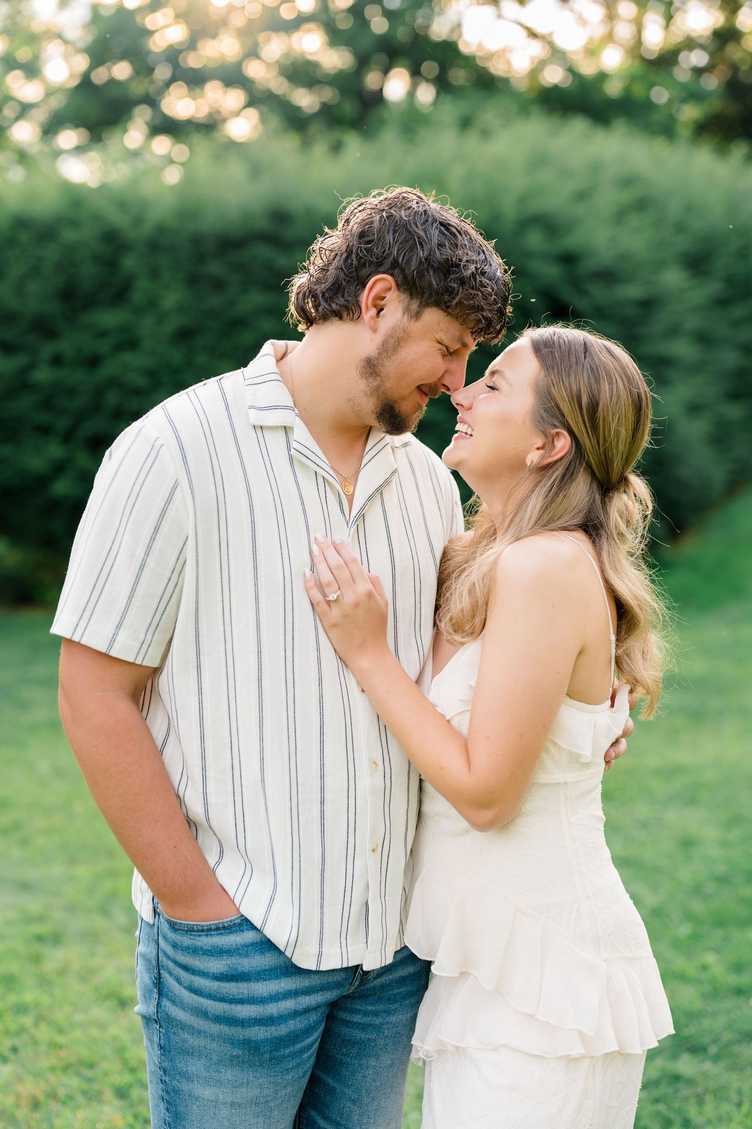 A couple shares a romantic moment outdoors, smiling at each other in golden hour light, surrounded by lush green hedges.