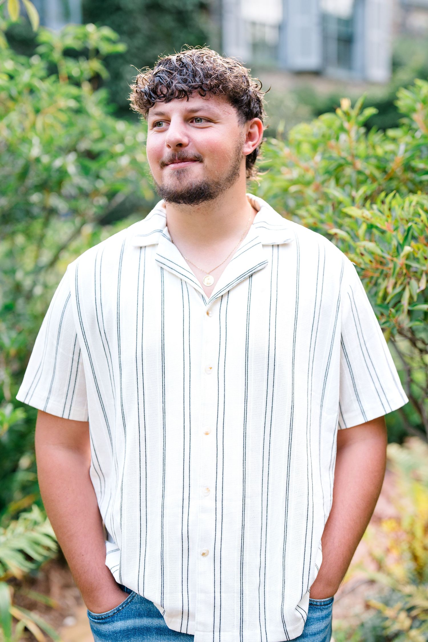 Young man in striped shirt standing outdoors with hands in pockets, smiling.