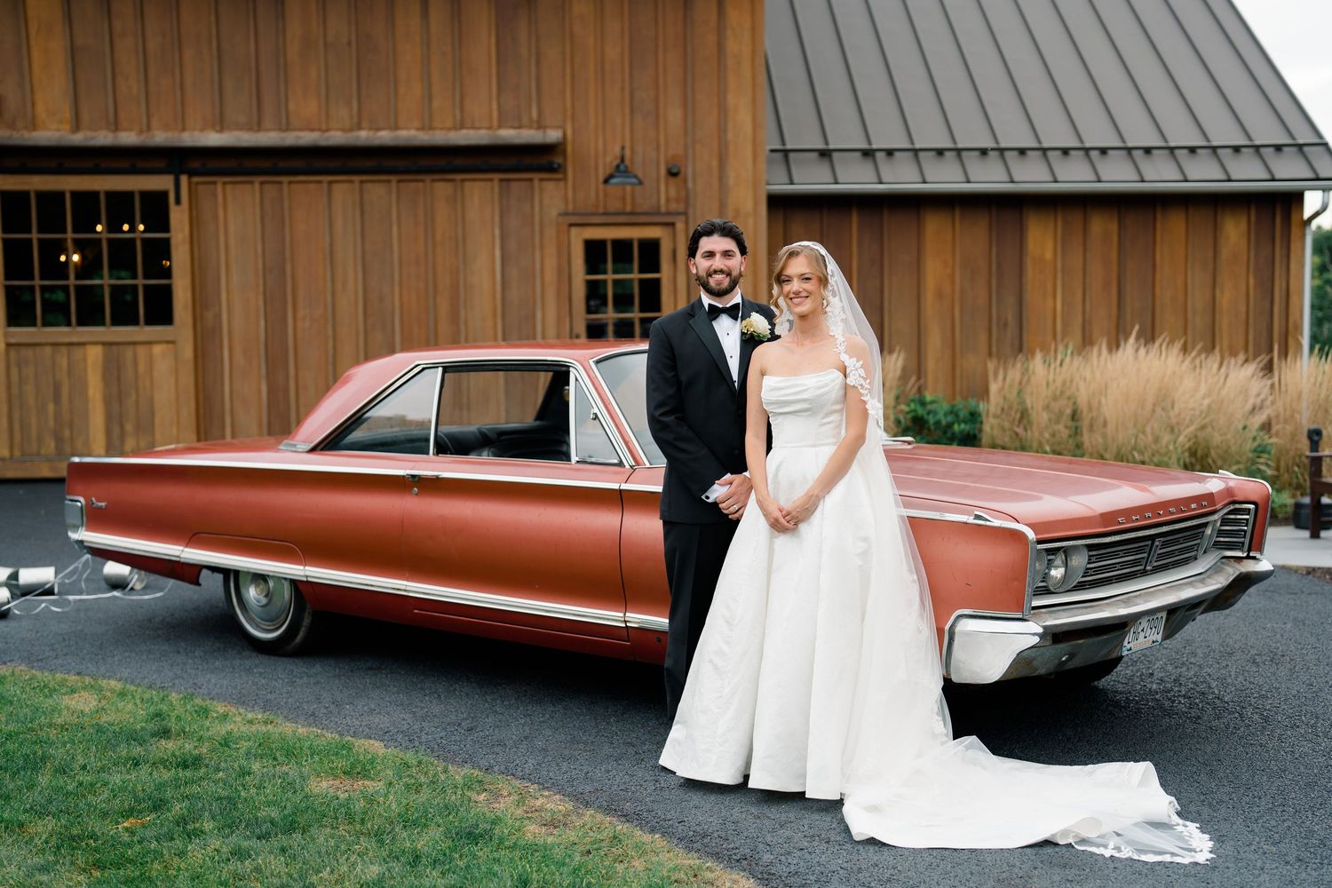Bride and groom posing beside a classic red vintage car outside a rustic barn wedding venue.