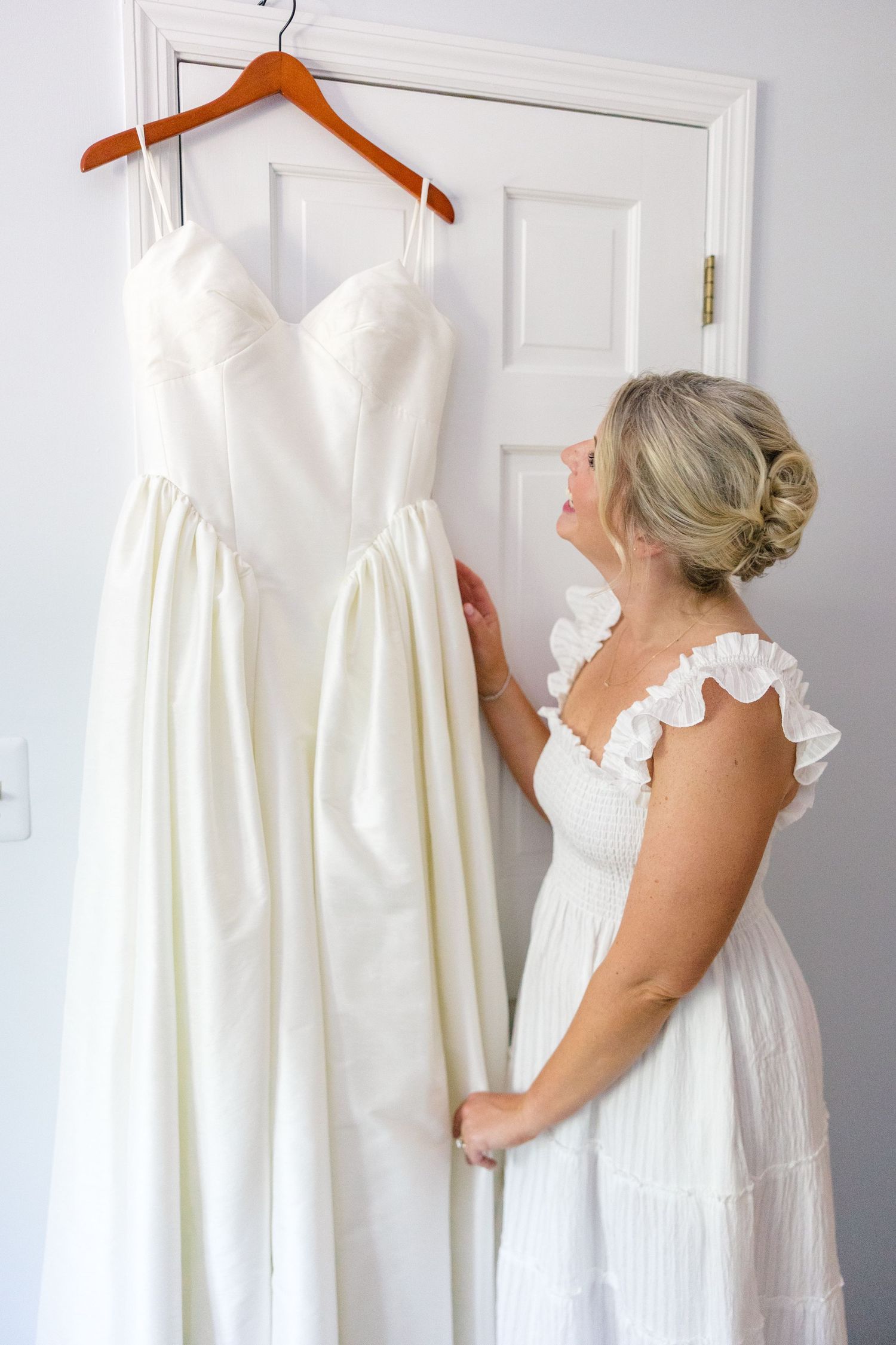 Bride admiring her white spaghetti strap wedding gown hanging on a wooden hanger on a white door.