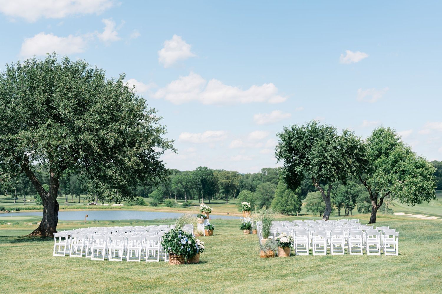 Outdoor wedding ceremony setup with white chairs on lush green lawn, surrounded by trees with a serene pond in the background.