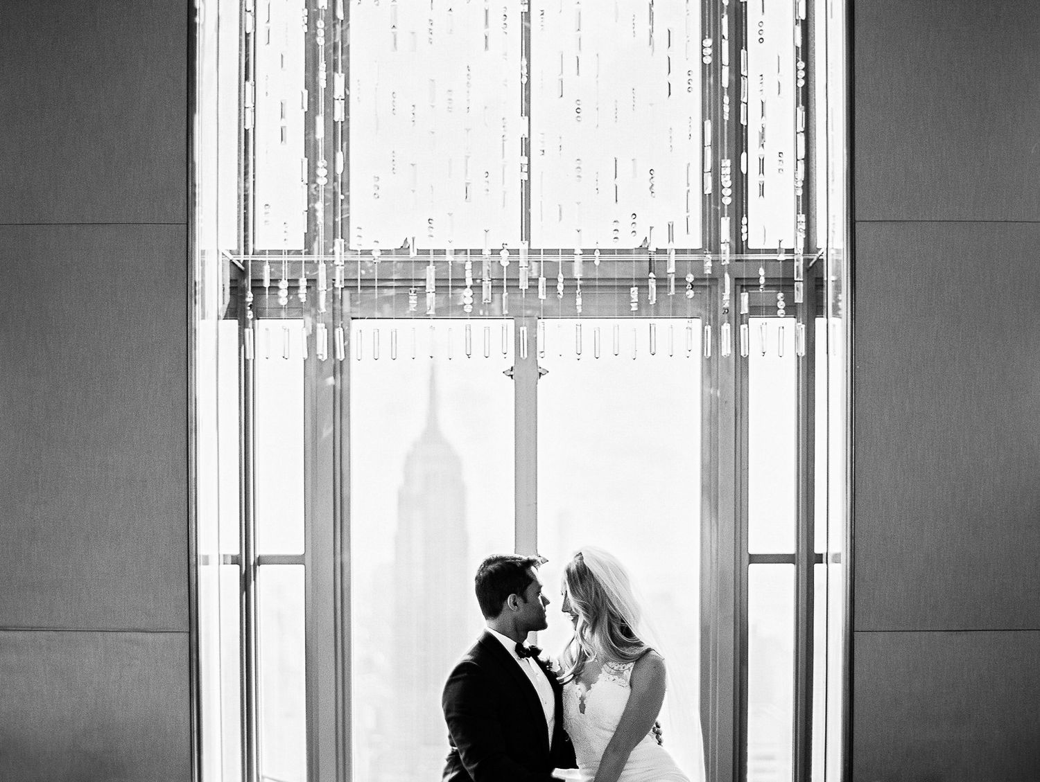 Black and white photo of a romantic couple embracing near a window with the Empire State Building visible in background.