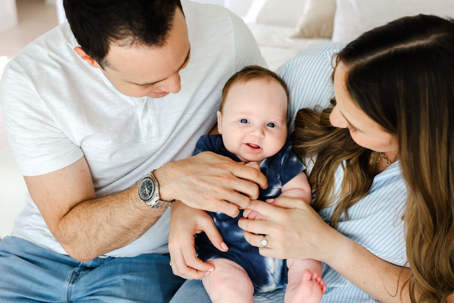 Loving parents playing with their small infant baby wearing a navy outfit while relaxing on bed together.