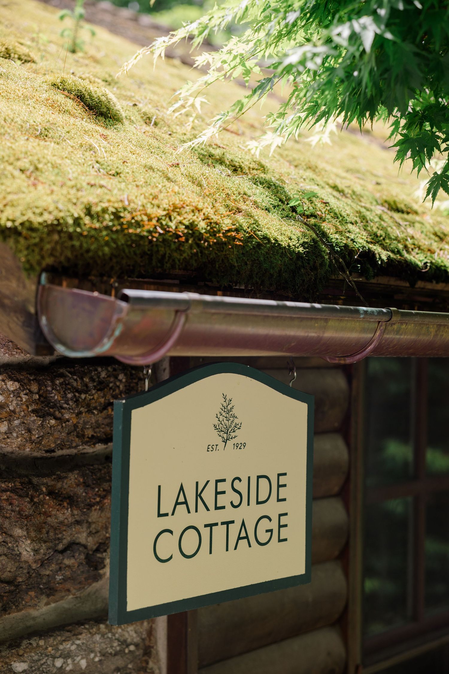 Rustic lakeside cottage sign mounted on stone wall with moss-covered gutter and overhanging foliage.