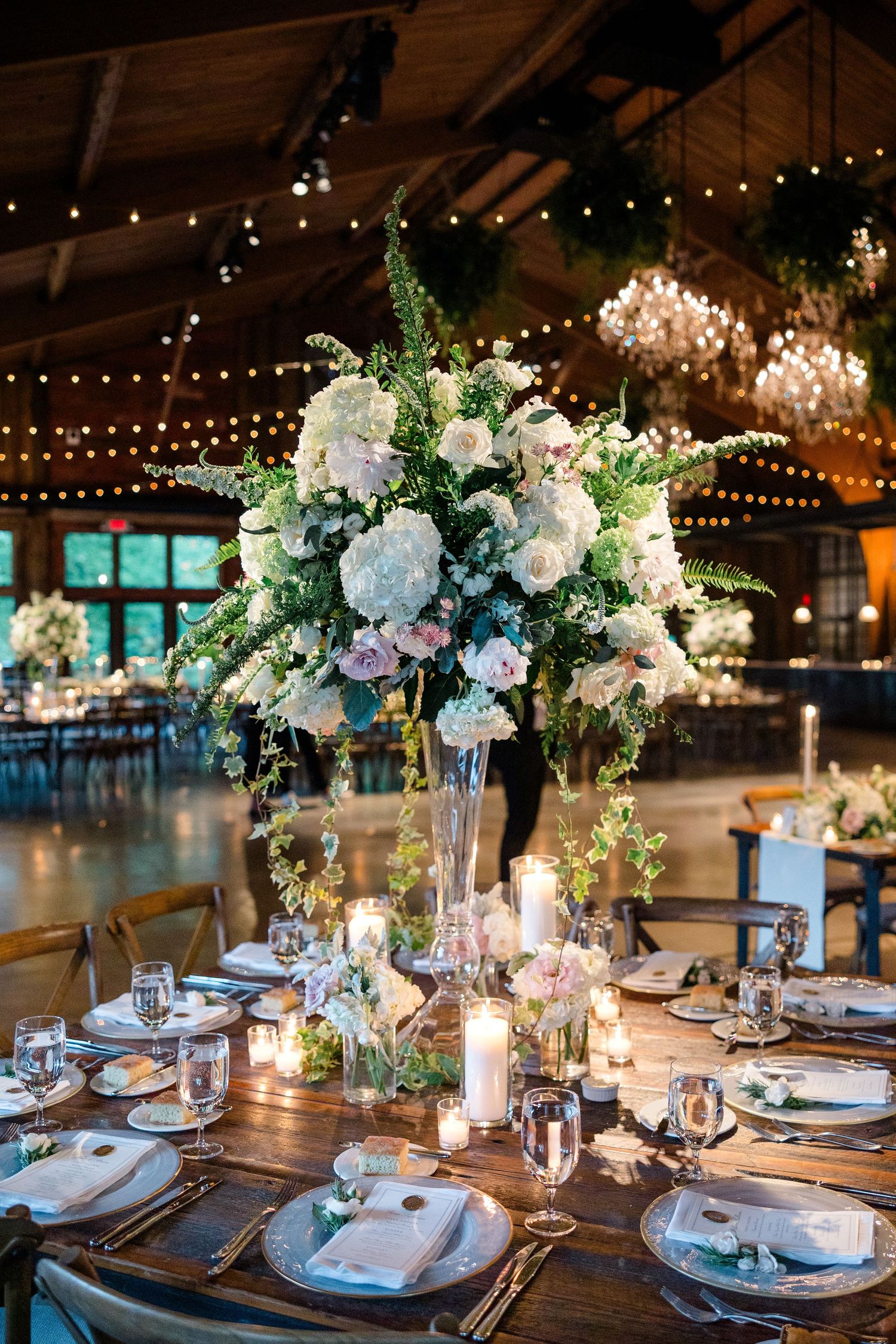 Elegant wedding reception table with tall white floral centerpiece and candles under string lights.