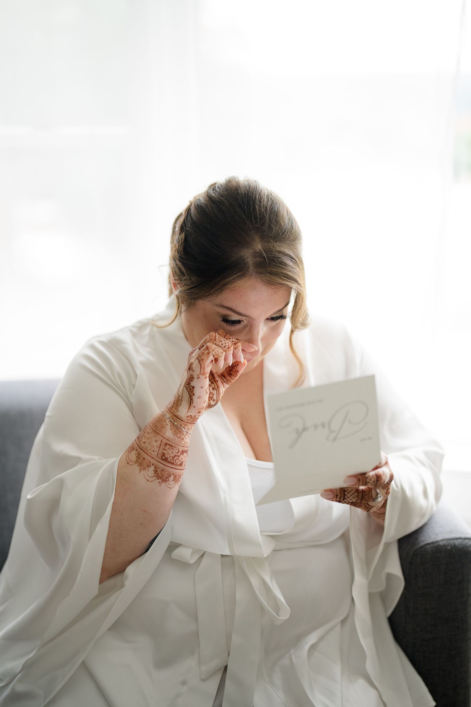 A person in white robe reads a card while sitting on couch with decorative henna on their hands.