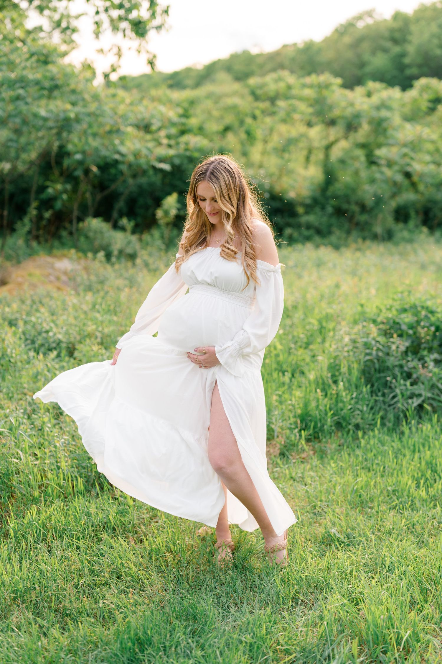 White dress flowing in the breeze in a sunlit meadow with green grass and trees.