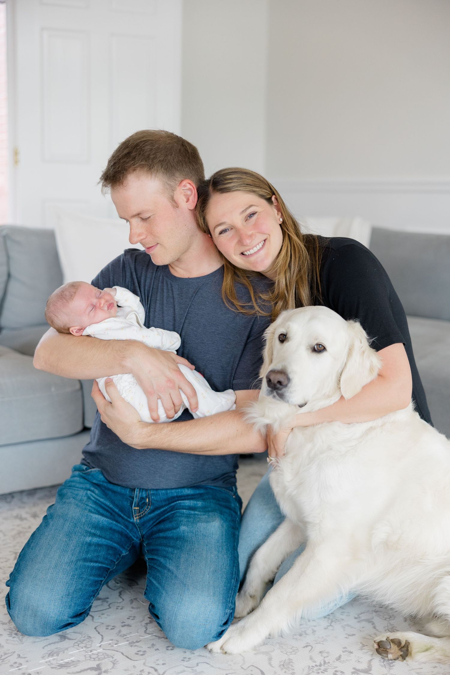 Family photo shoot on grey carpet with white dog and newborn baby in casual home setting.