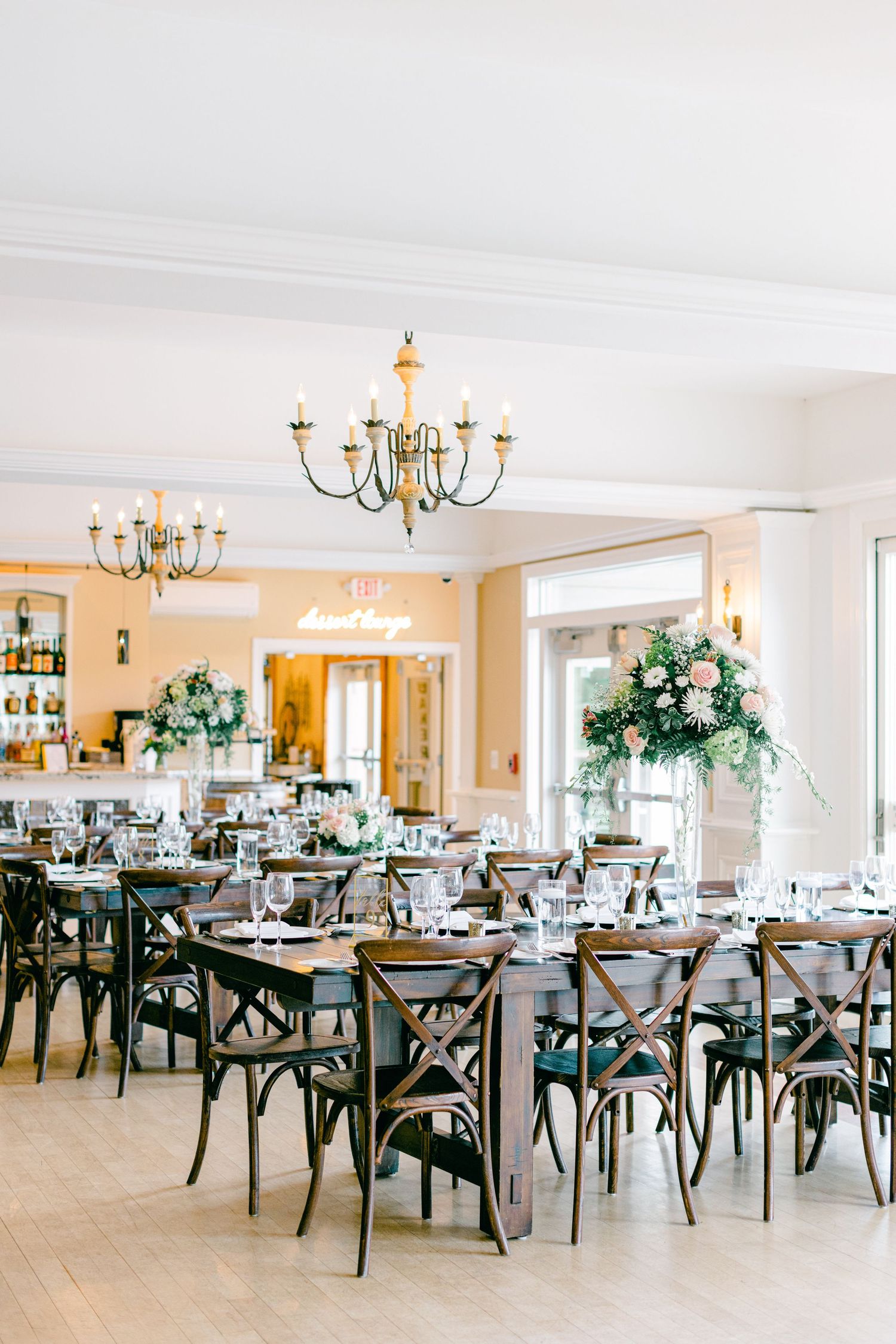 Elegant wedding reception venue setup with rustic cross-back chairs and brass chandeliers in a bright white ballroom.