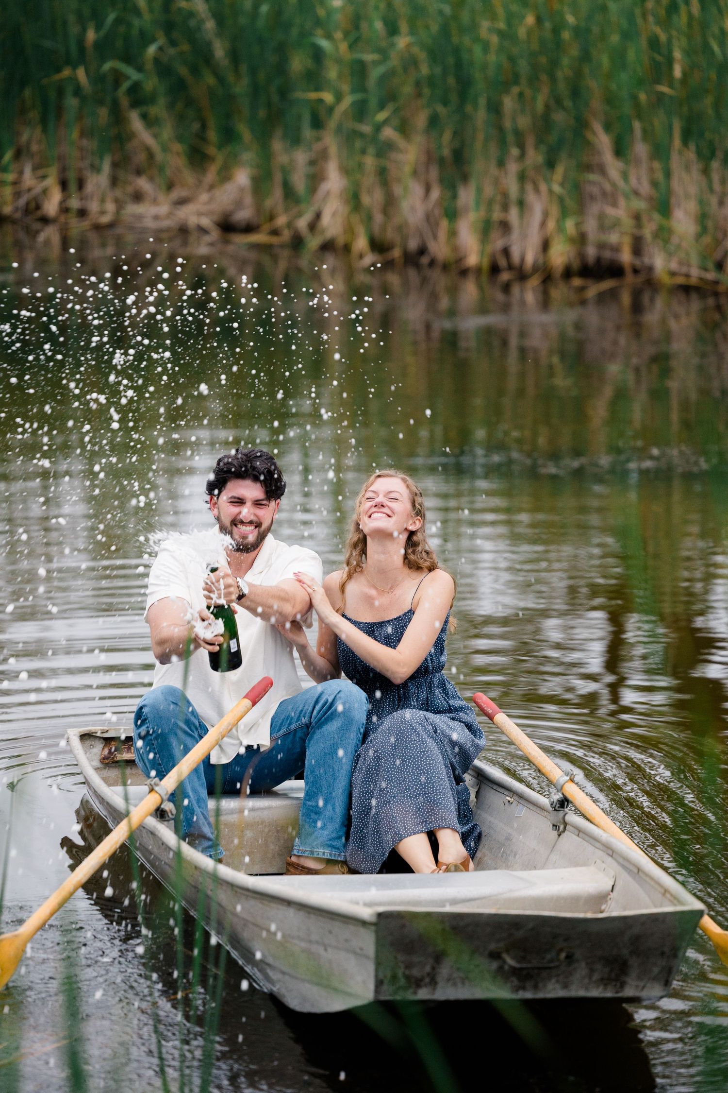 Two people sharing joyful laughter while sitting in a small boat on a tranquil lake surrounded by reeds.