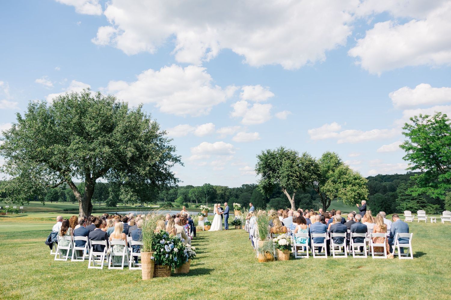 Outdoor wedding ceremony on green lawn with white chairs, large trees, and blue sky with clouds.