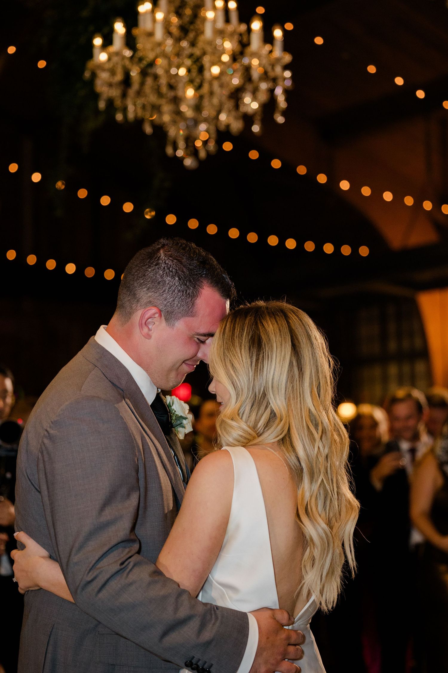 A romantic couple shares a dance under a crystal chandelier with warm string lights glowing overhead.