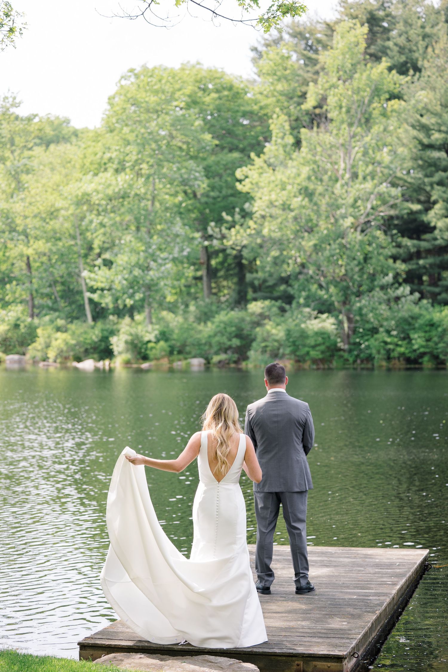 A couple stands on a wooden dock by a serene lake surrounded by lush green trees.