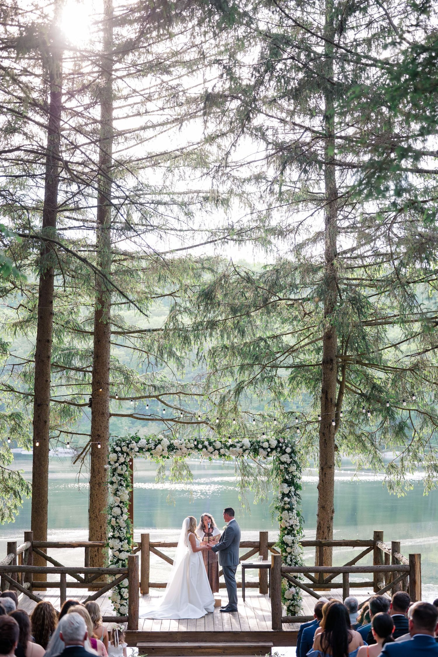 Outdoor wedding ceremony under tall pine trees with a stunning lake backdrop and floral arch.
