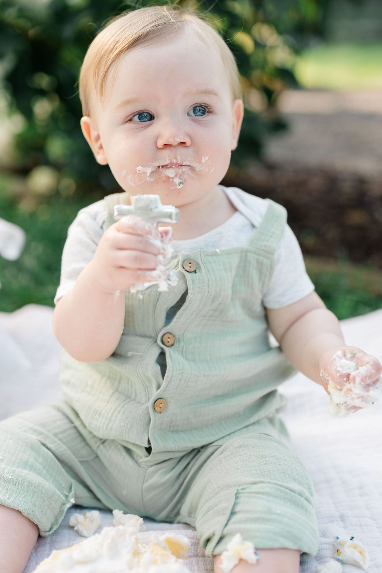 Cute baby boy in green romper eating smash cake with frosting on his face outdoors.
