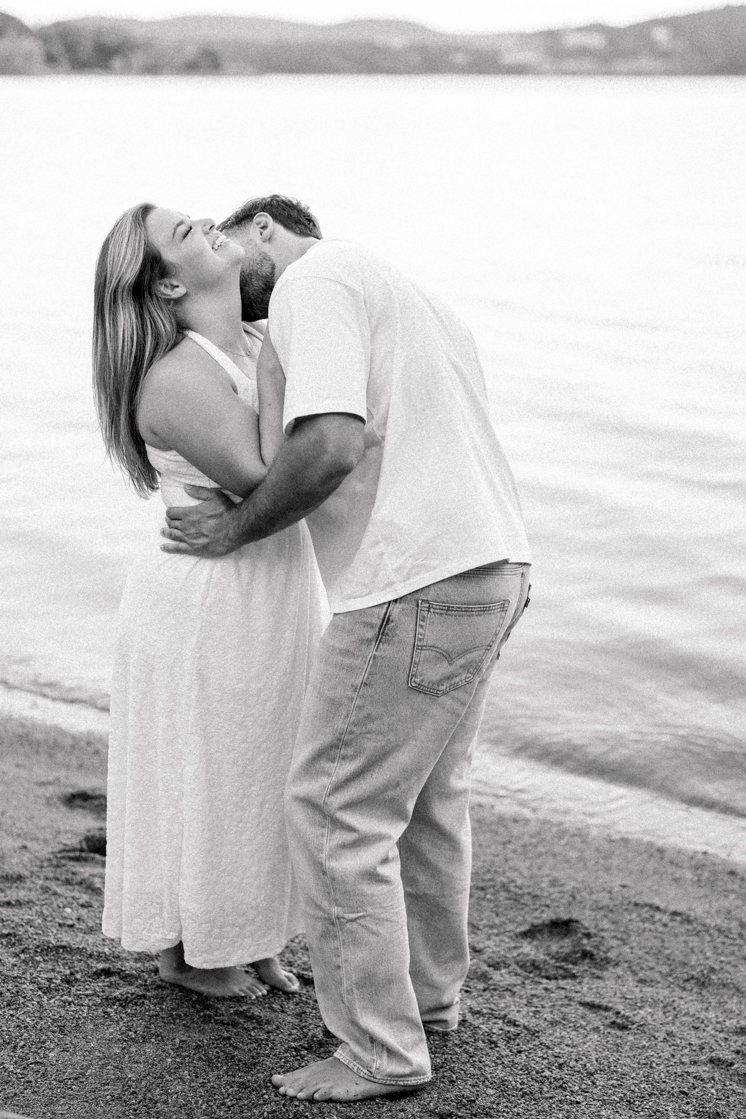 Black and white photo of a couple kissing barefoot on a sandy beach shore with calm water in the background.