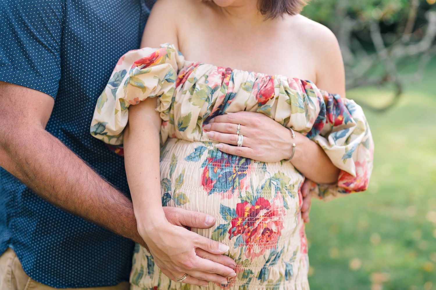 Pregnant woman in floral off-shoulder dress cradling baby bump with partner outdoors.