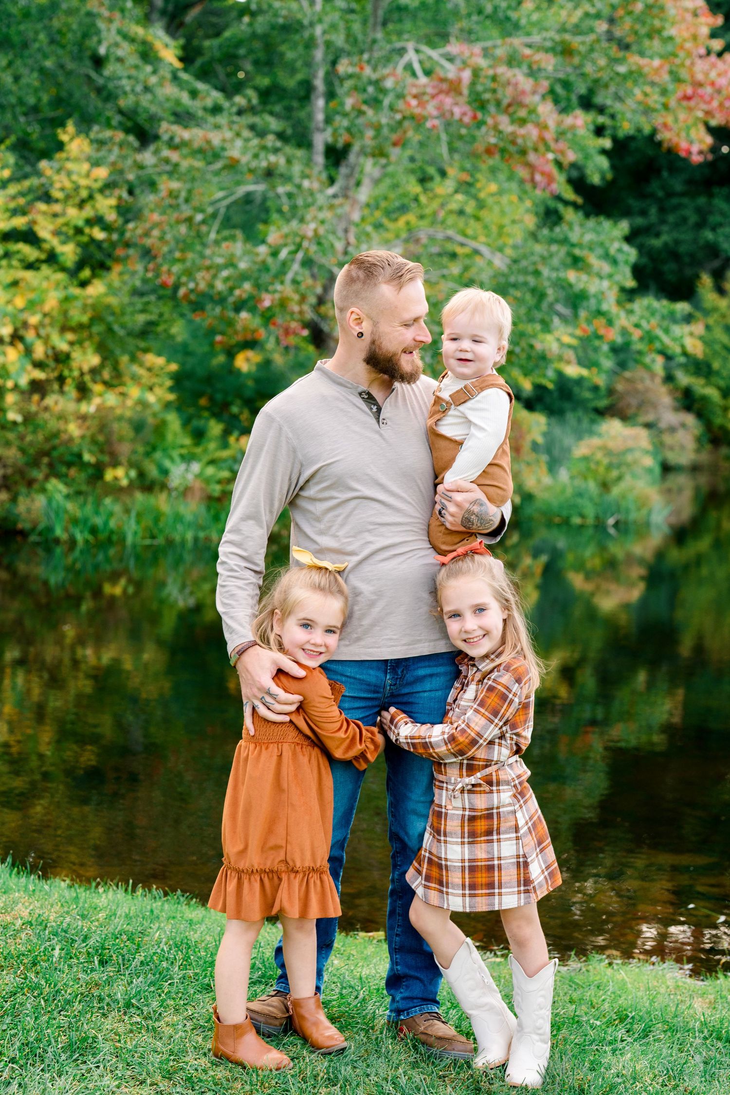 A bearded father holds a baby while two young girls in fall dresses hug him outdoors near a pond.