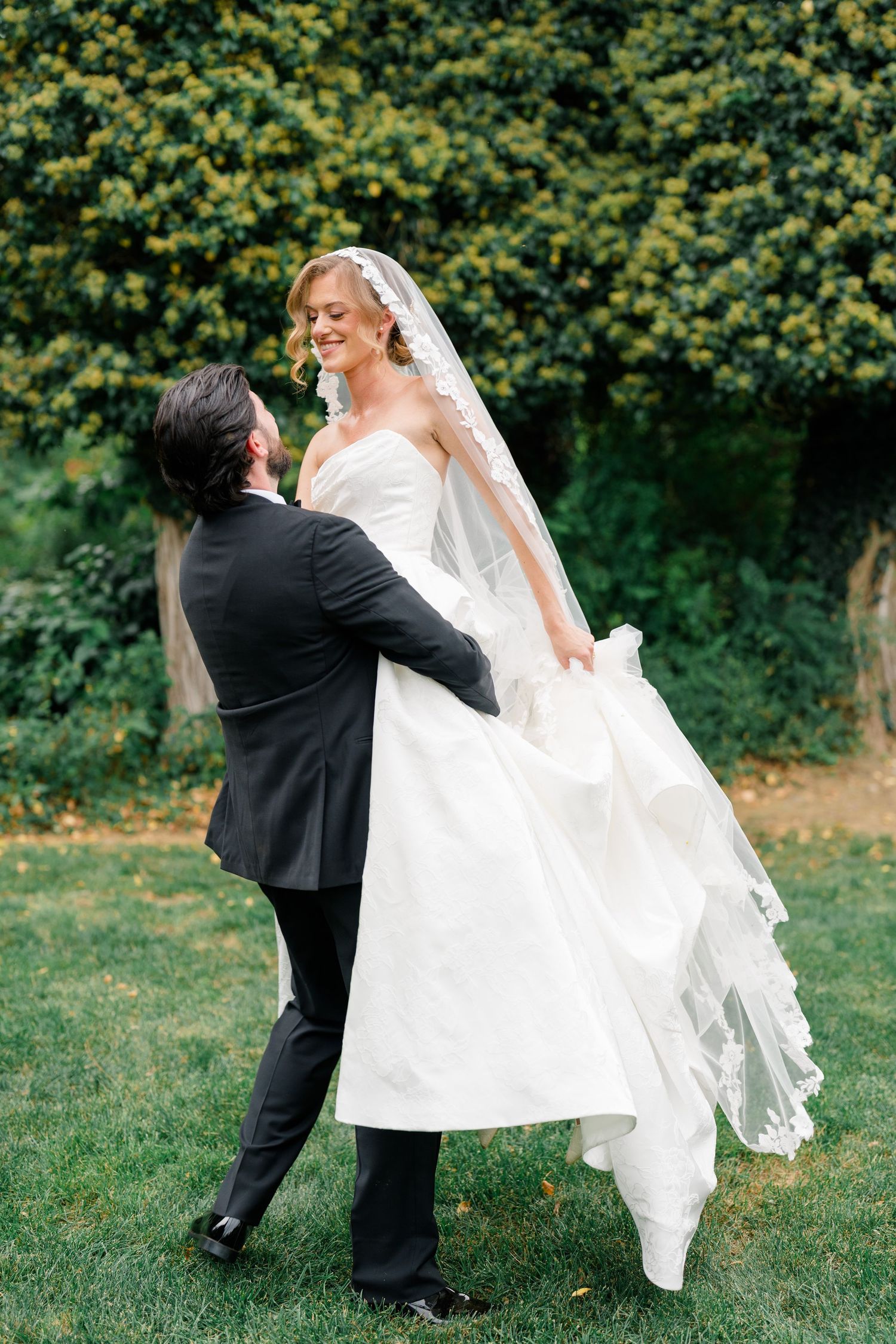 Groom lifts smiling bride in white lace gown and cathedral veil in lush green garden setting.