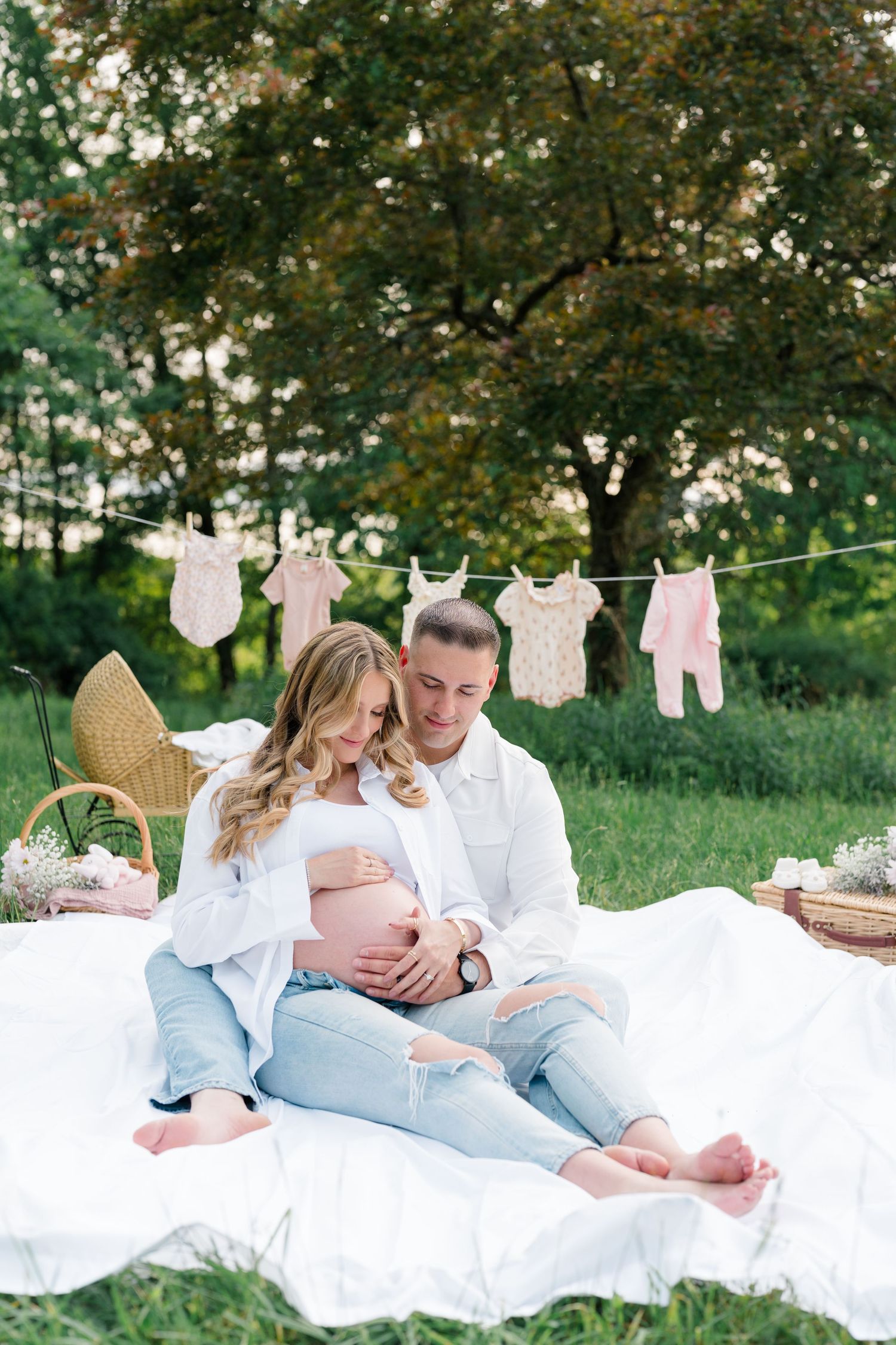 Expecting couple sits on white blanket outdoors with baby clothes hanging on clothesline and picnic basket nearby.