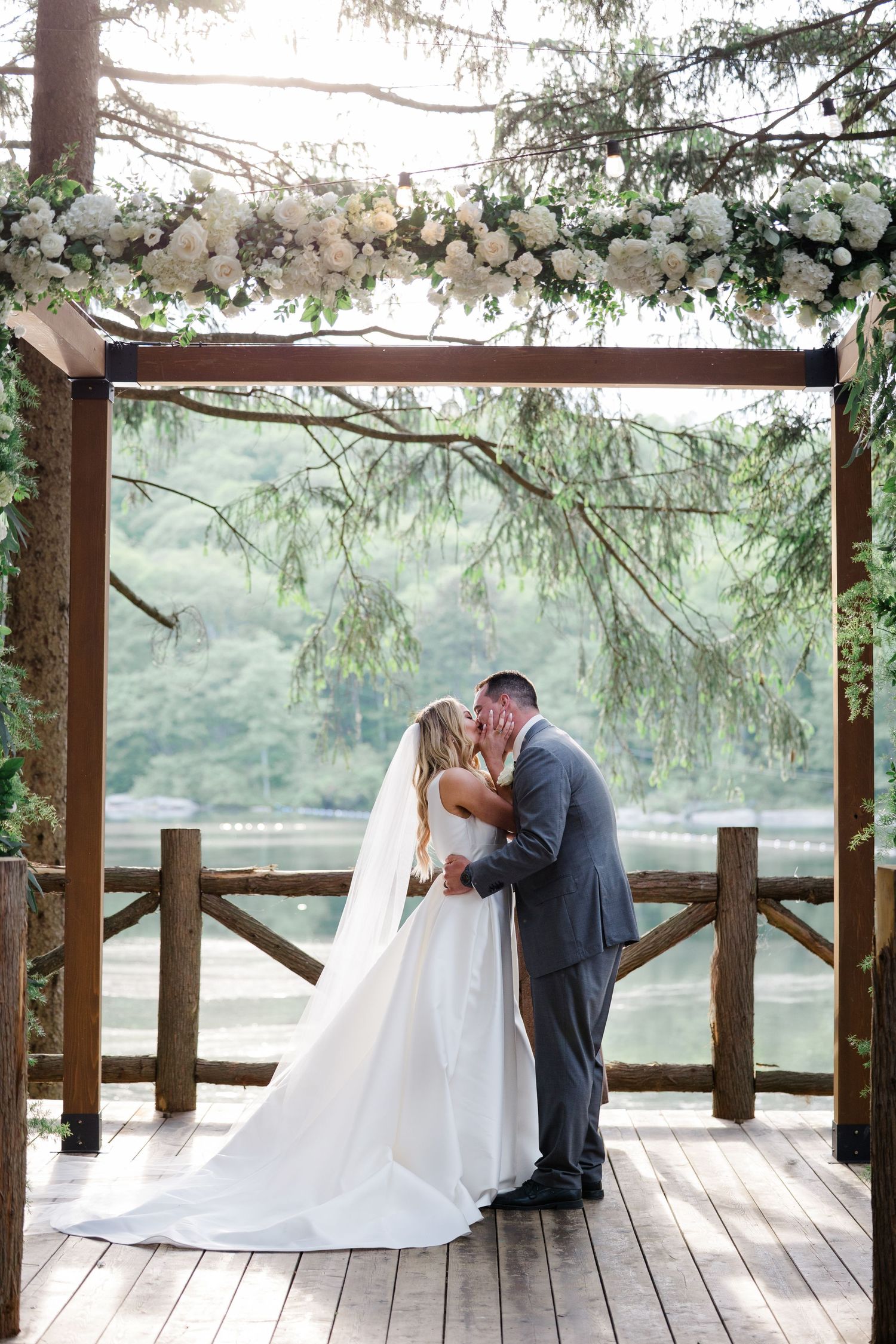 A romantic wedding ceremony takes place on a wooden deck overlooking mountains, with floral decorations on a wooden arch.
