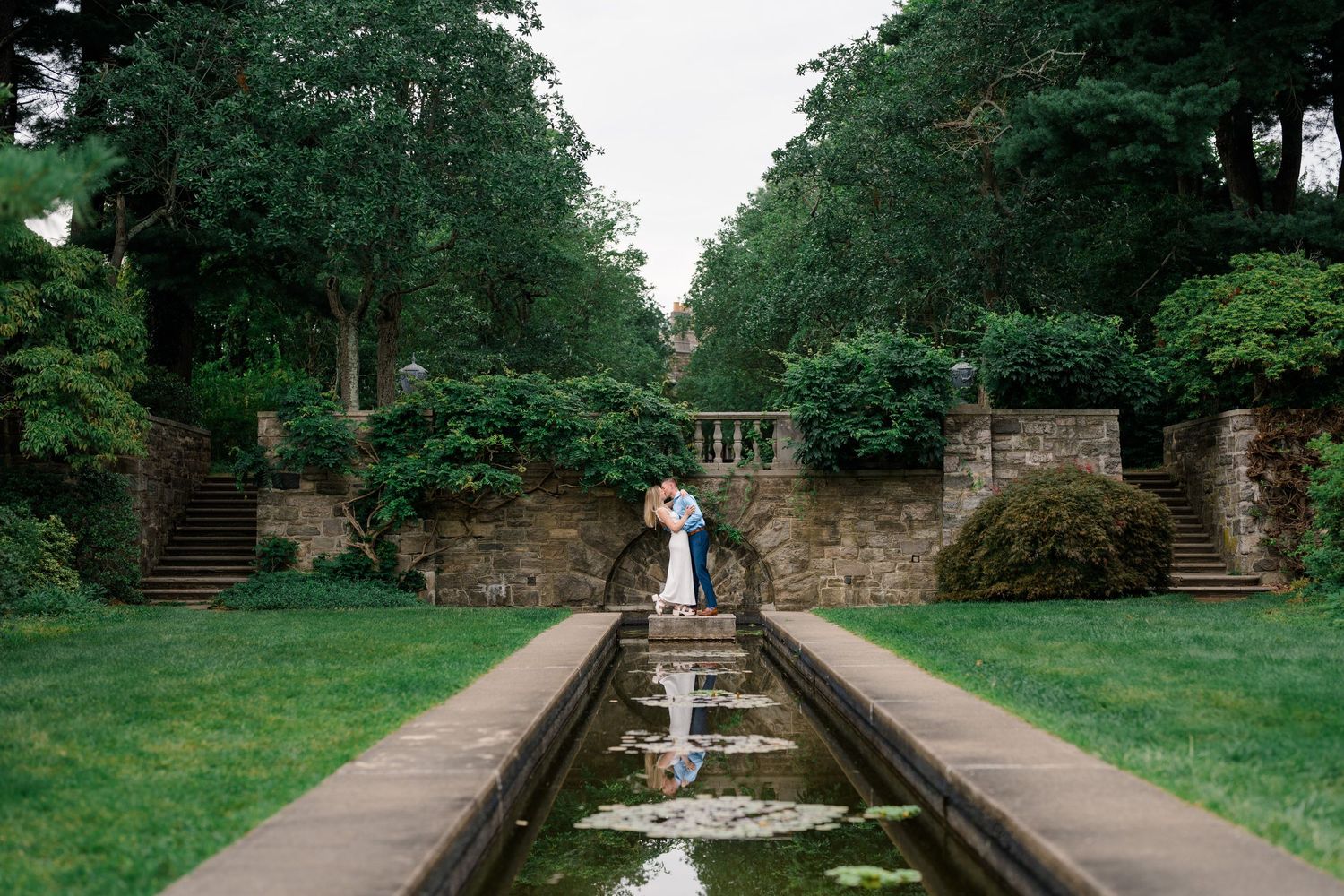 Couple sharing a romantic kiss beside a reflective garden canal surrounded by lush greenery and stone walls.
