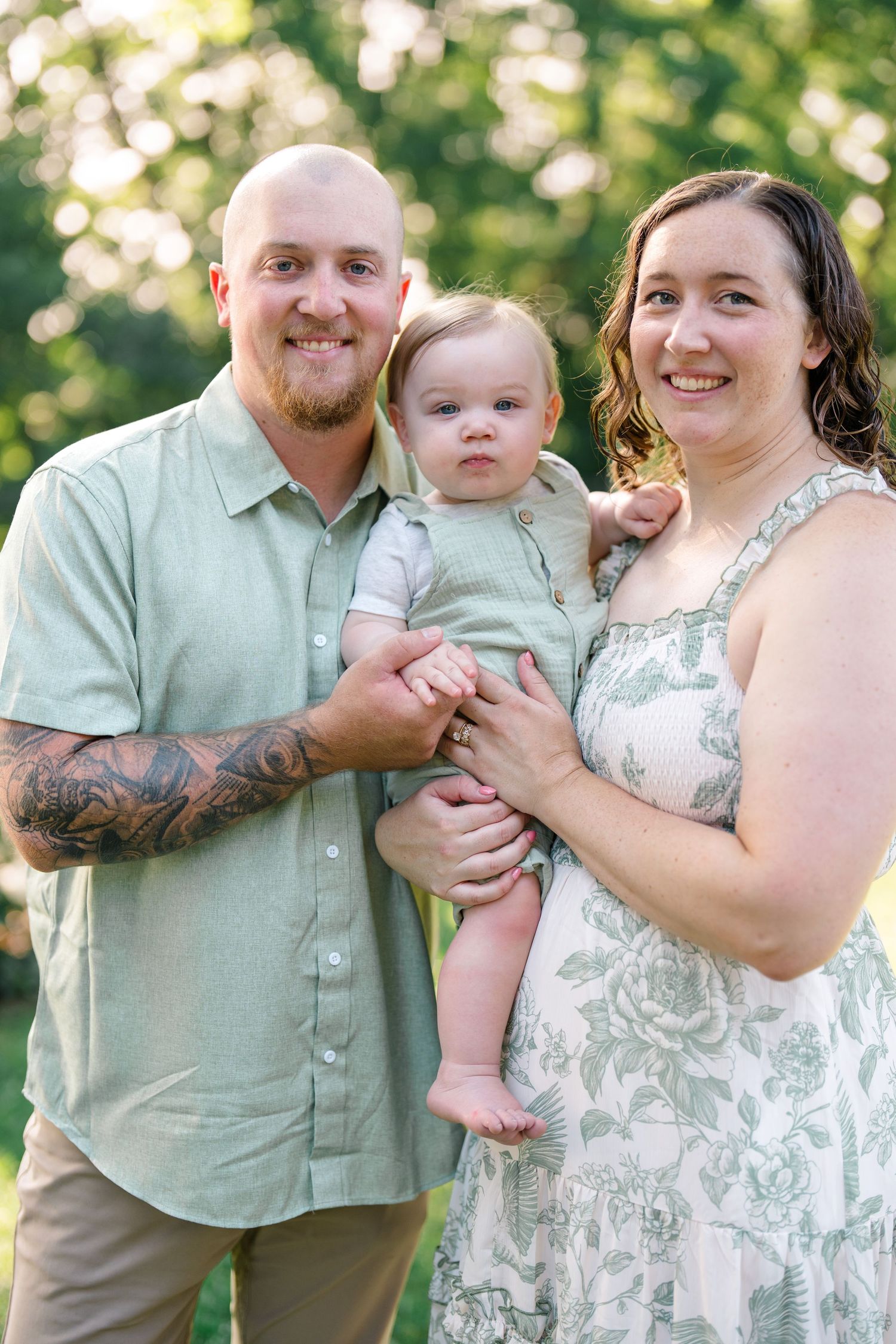 Happy family of three with tattooed dad, mom in floral dress, and baby in green romper outdoors.