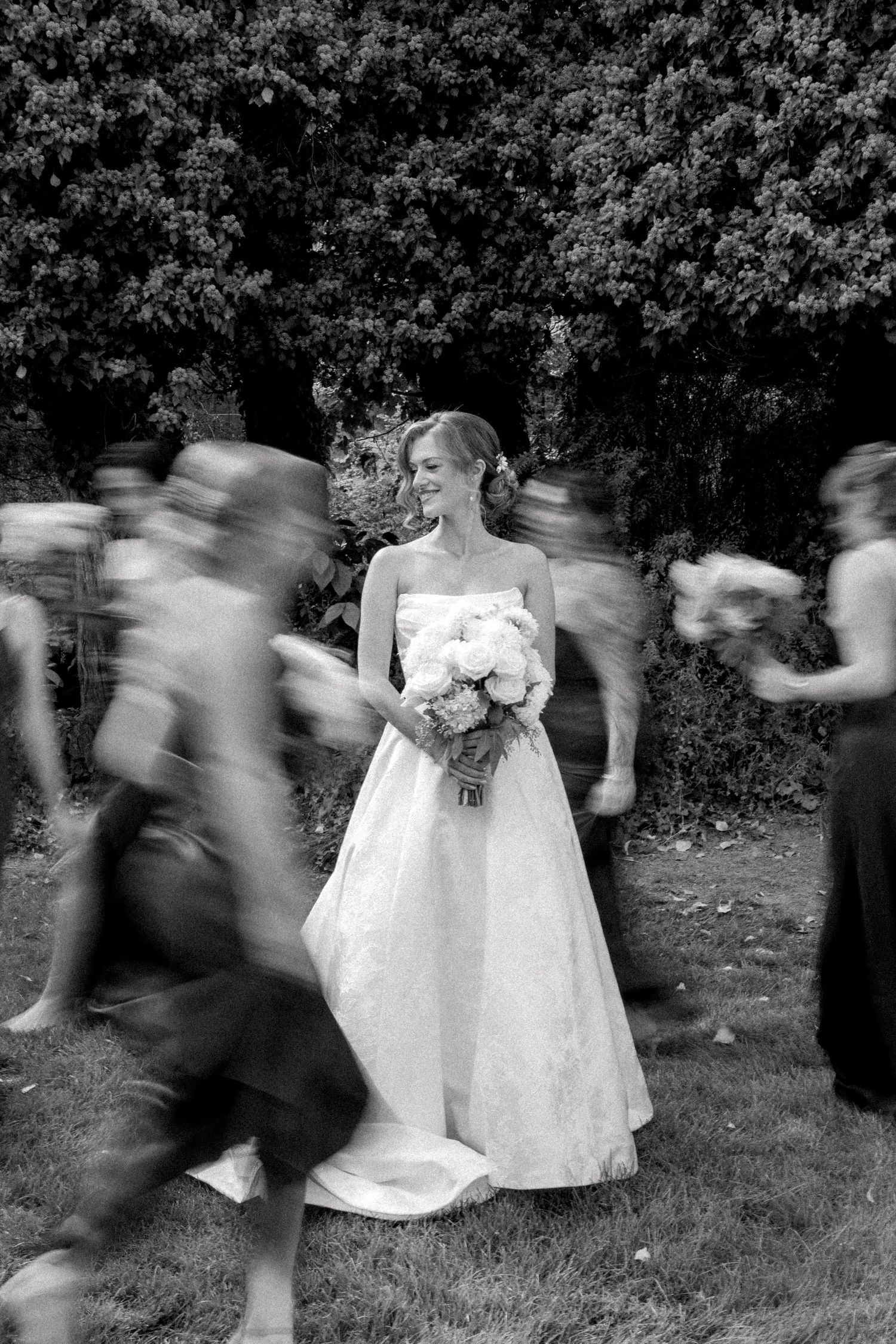 Black and white photo of a smiling bride in a white gown holding a bouquet, surrounded by blurred moving guests outdoors.
