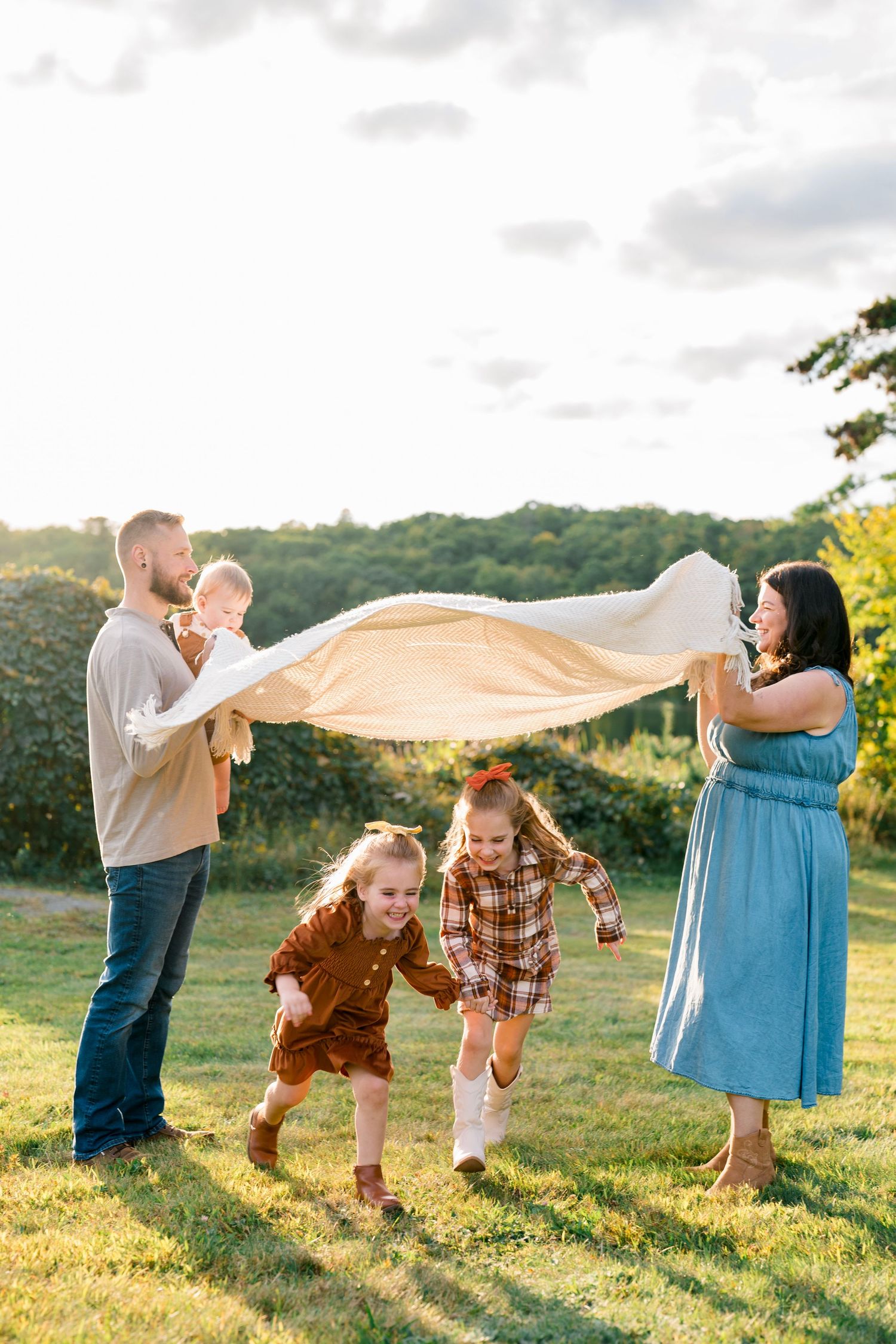 Happy family playing outdoors with a blanket on a sunny day, two girls running while parents hold a baby.