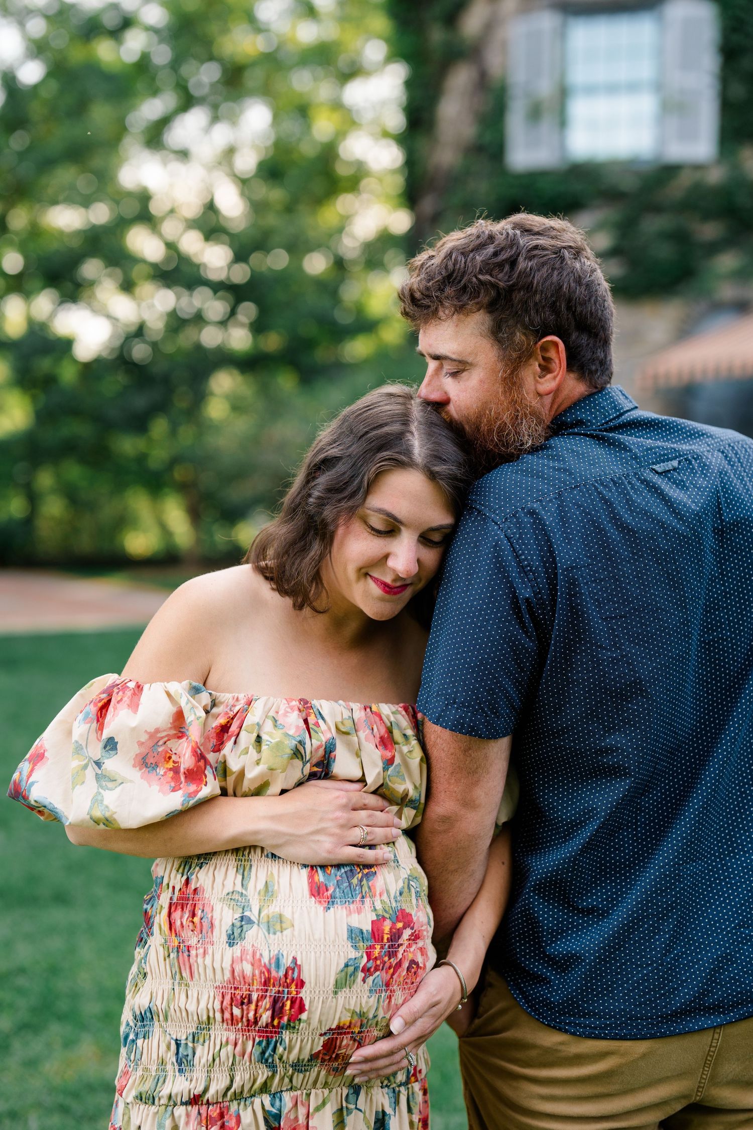 Couple sharing a tender moment outdoors, man kissing pregnant woman's head while embracing her baby bump in a floral dress.