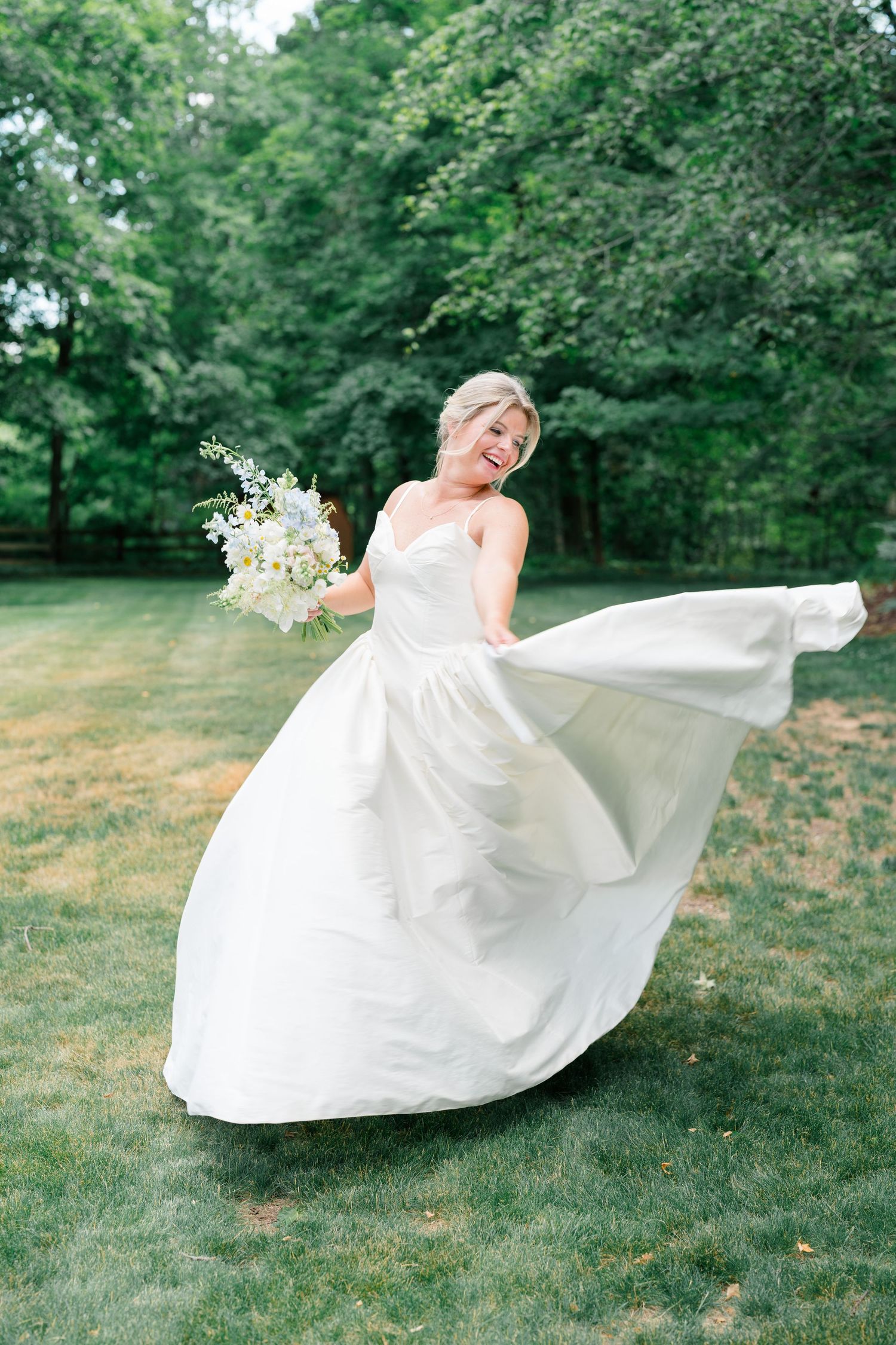 Joyful bride twirls in flowing white satin wedding gown while holding white floral bouquet on green lawn.