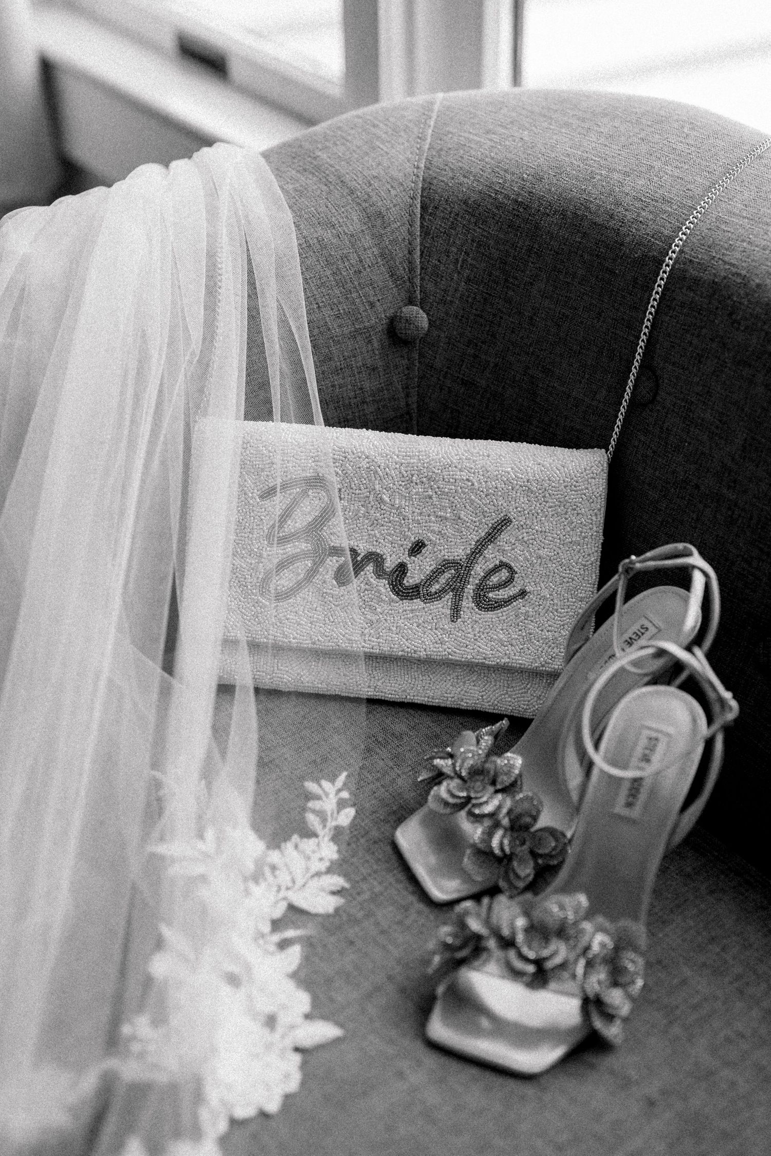 Black and white wedding detail photograph showing a bridal veil, embroidered 'Bride' sign, and elegant heels on a tufted chair.