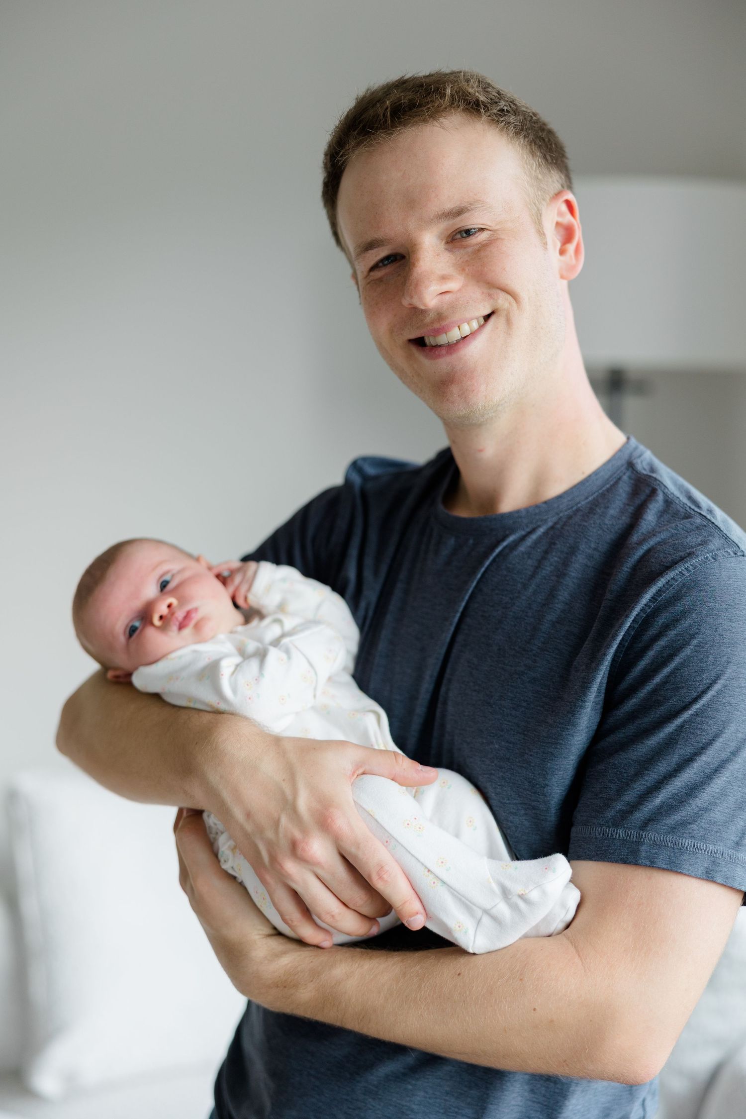 A person in a dark blue t-shirt holds and smiles at their newborn baby wrapped in white.