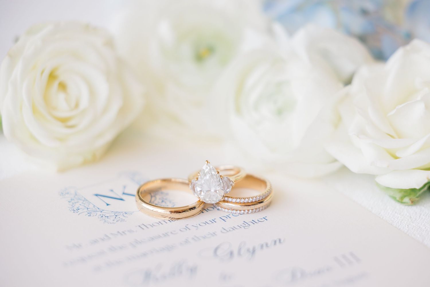 Gold wedding bands and pear-shaped diamond engagement ring displayed on elegant wedding stationery with white roses.